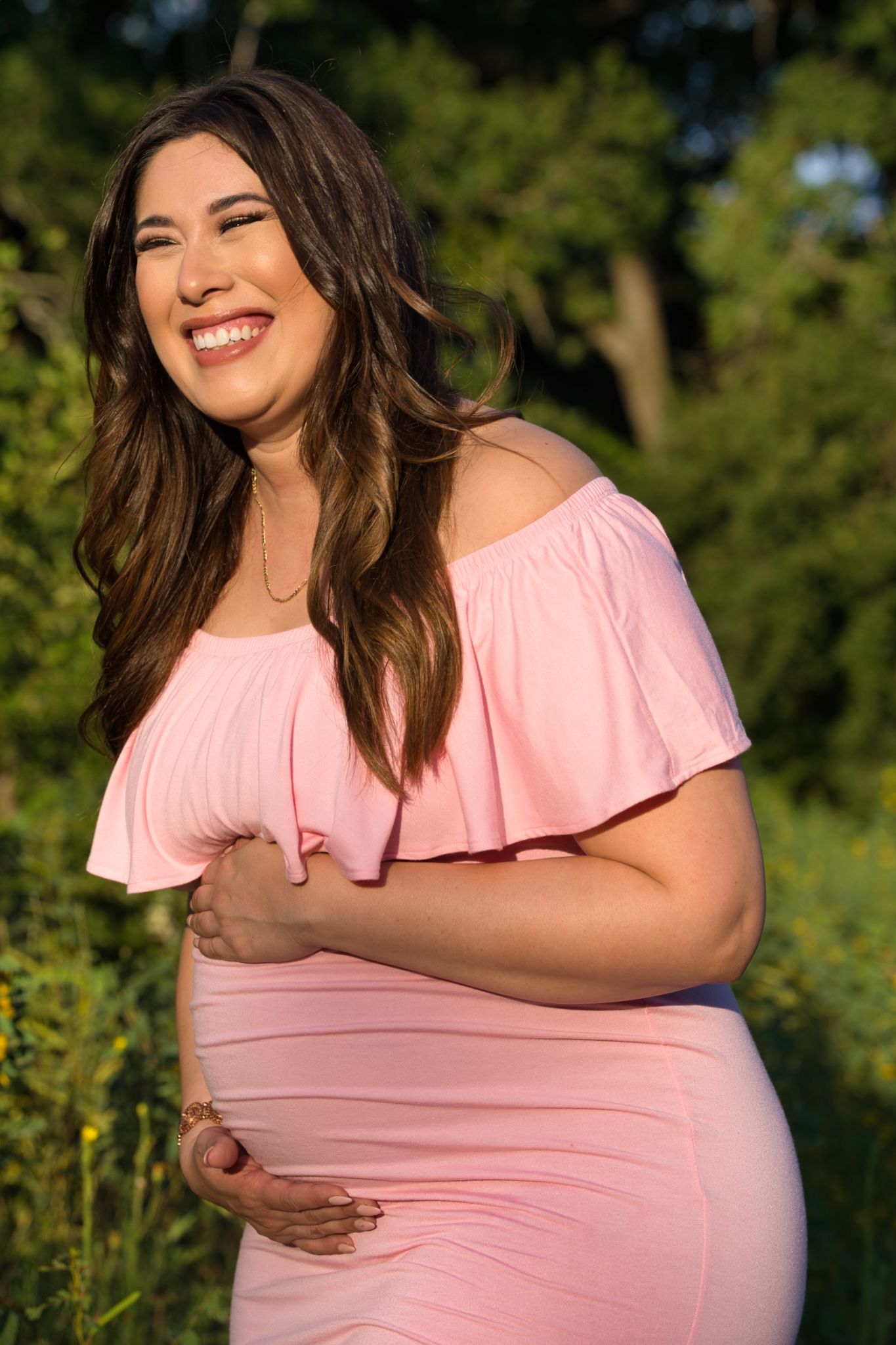Expecting mother in pink off-shoulder dress smiles outdoors in natural setting.