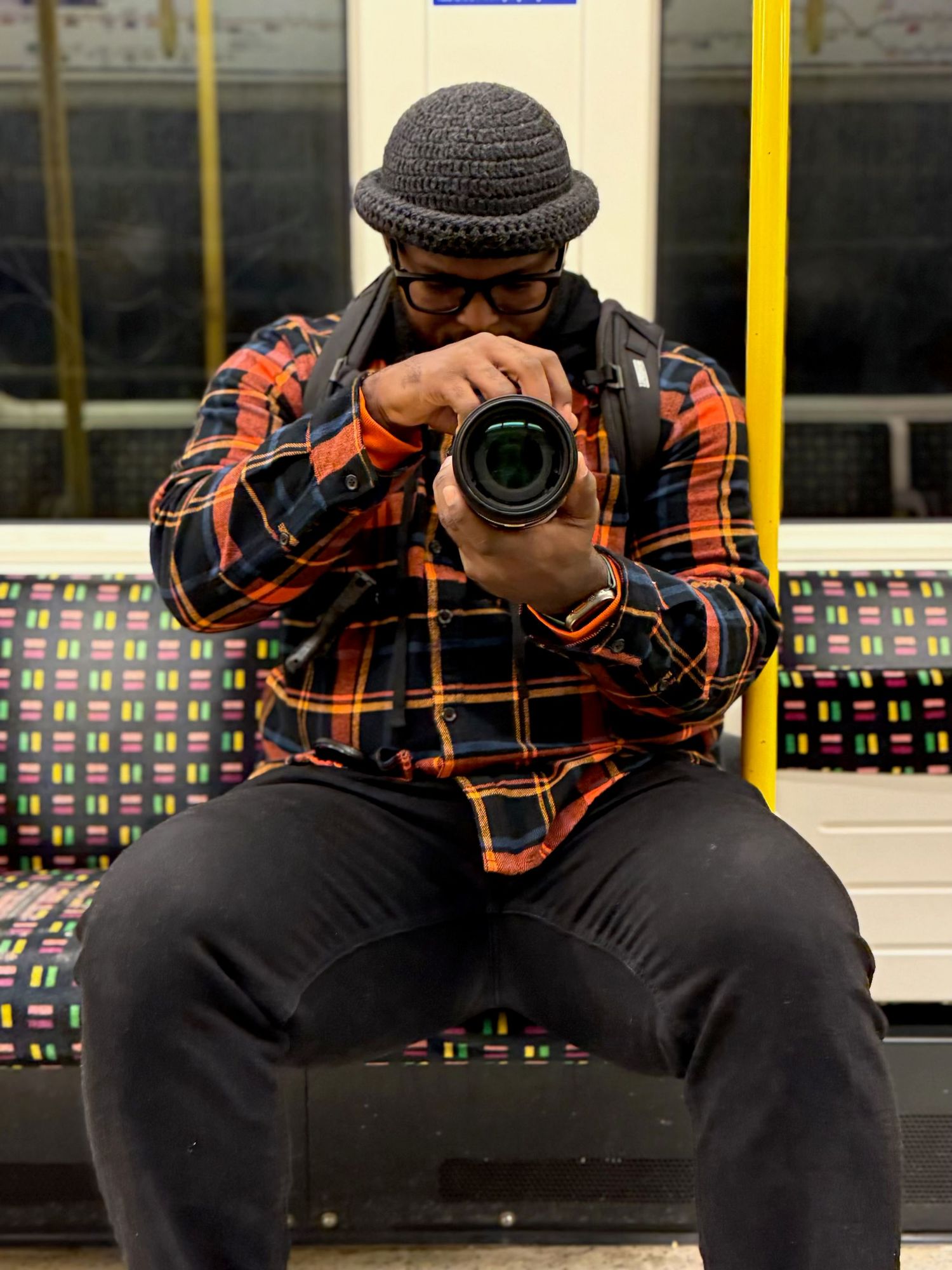 Photographer in plaid shirt and bowler hat holding camera while sitting on subway train.