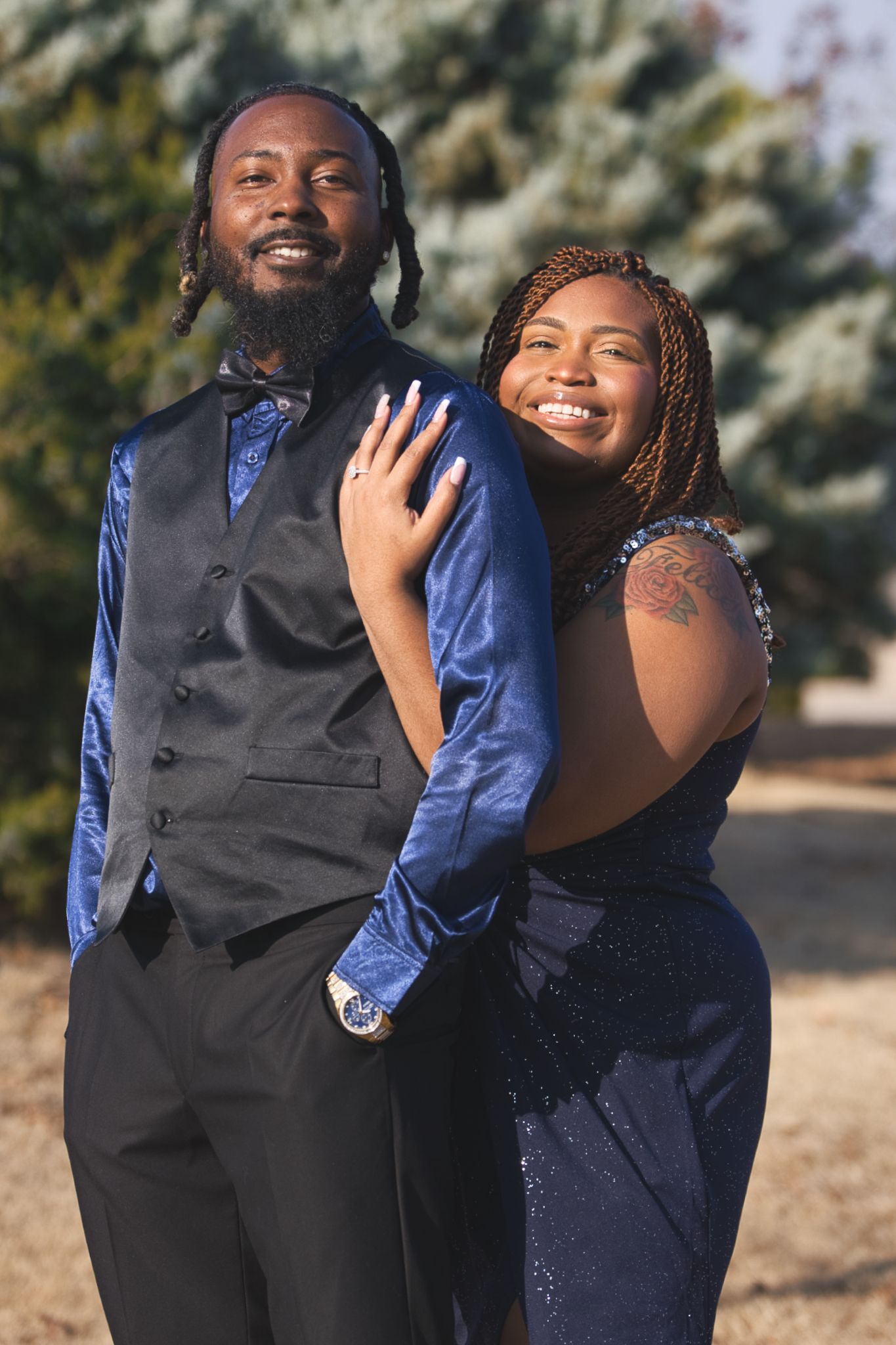 A couple in formal navy blue attire smiling together outdoors with evergreen trees in the background.