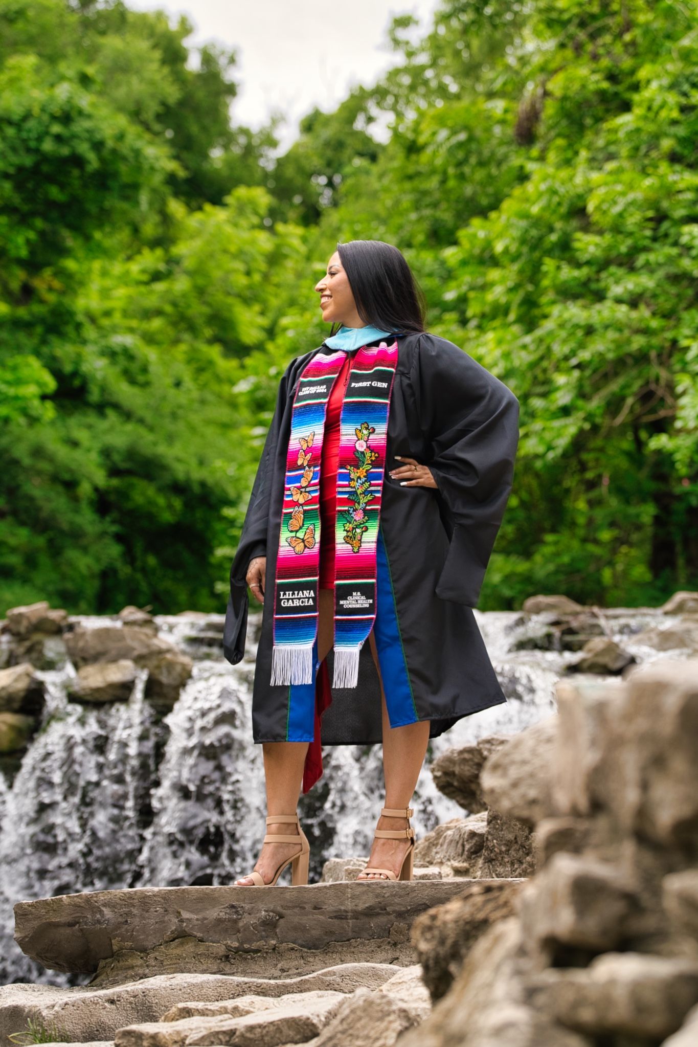 Graduate wearing a decorated stole stands near a waterfall in a lush green setting during a graduation photoshoot.