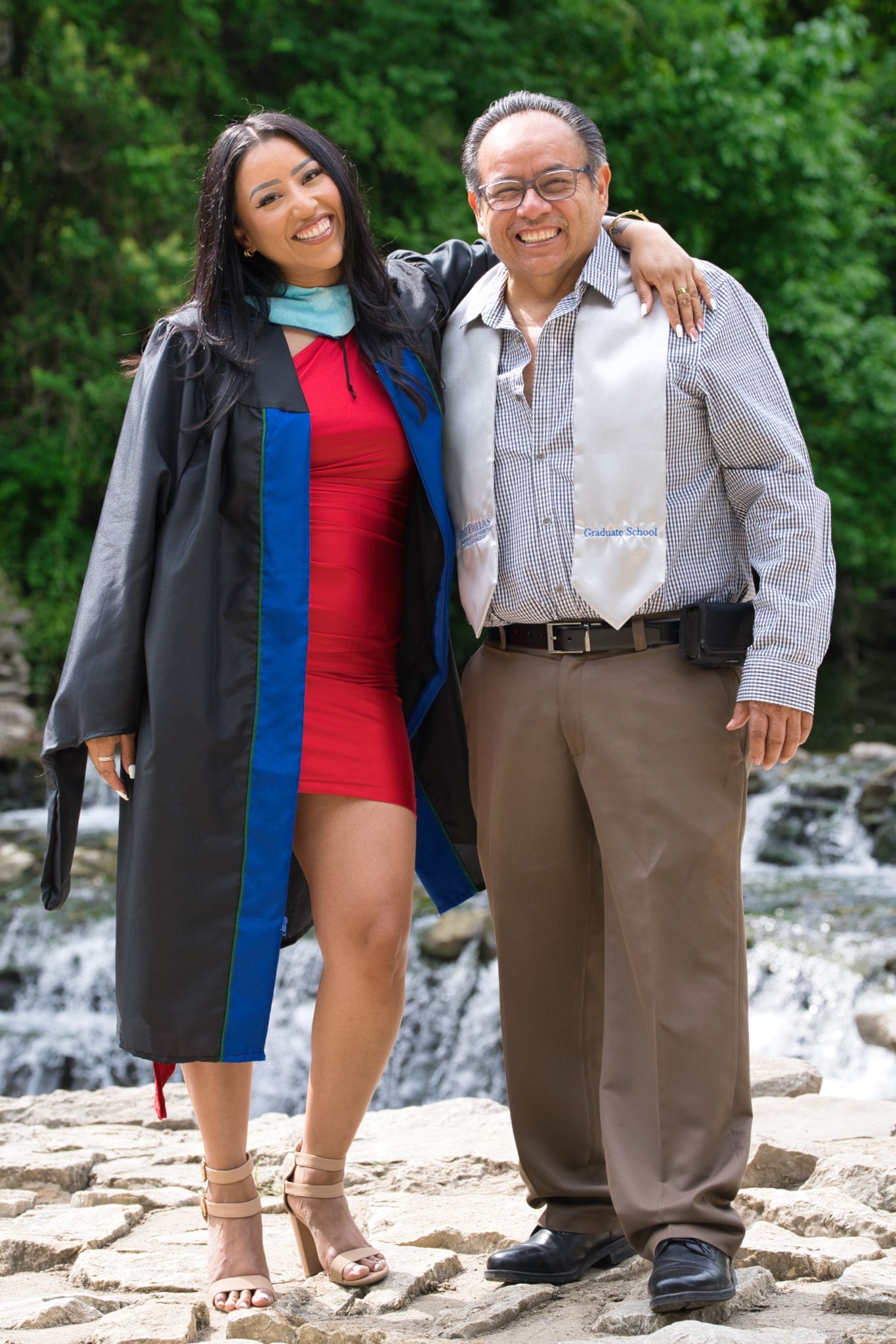 Graduate in red dress and academic regalia poses with family member by scenic waterfall.