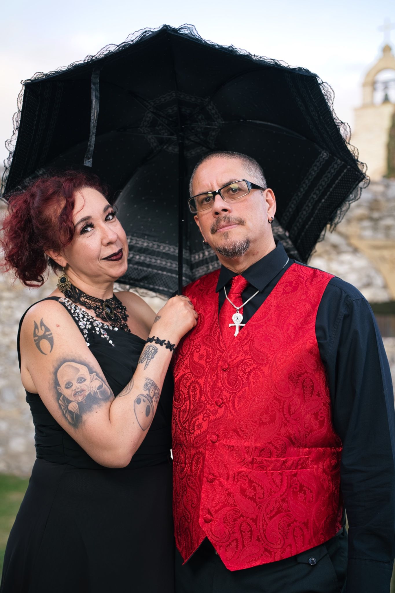 Gothic couple poses with black lace umbrella, wearing red brocade vest and black formal attire at stone building.