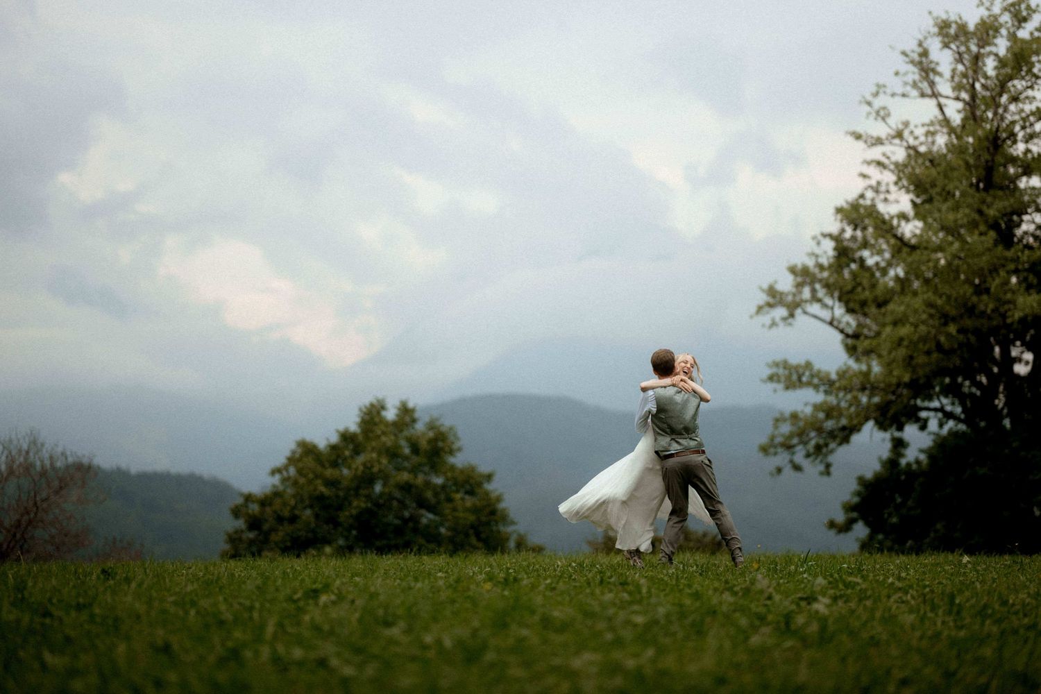 Jana & Lukas tanzen lachend auf einer grünen Wiese mit Bergpanorama in Vorarlberg – natürliche Hochzeitsfotografie voller Freude und Emotion