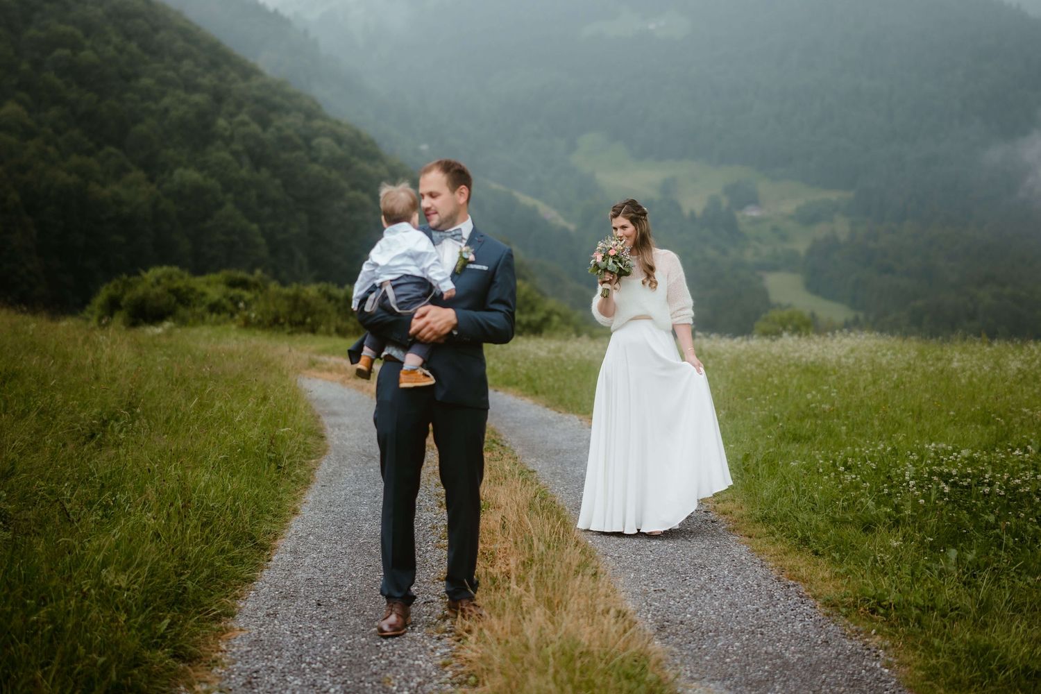 Stefan trägt ein kleines Kind auf dem Arm, während Jasmin mit ihrem Brautstrauß lächelnd folgt – Hochzeitsreportage in Vorarlberg