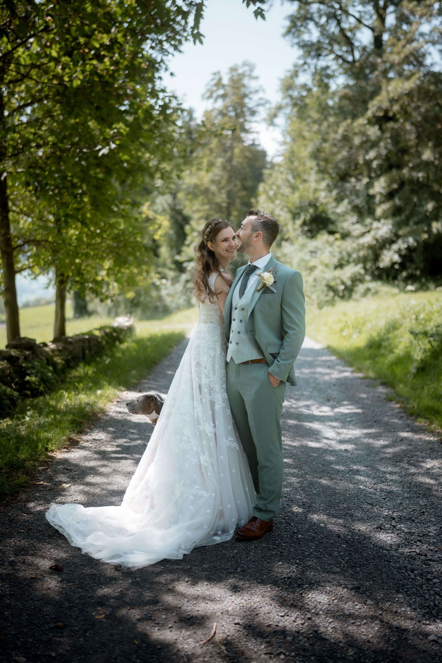 Anna und Markus posieren mit ihrem Hund auf einem sonnigen Waldweg – natürliche Hochzeitsfotos in Vorarlberg von NuMa Fotografie