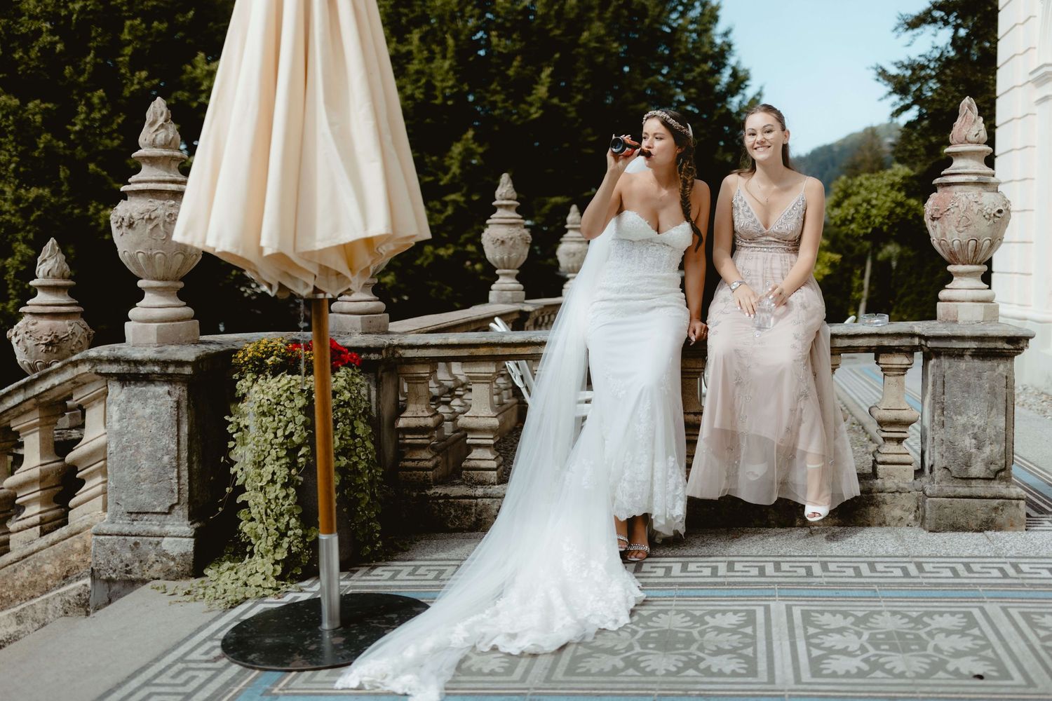 Lässige Braut mit Bierflasche und lachender Trauzeugin auf einer historischen Terrasse bei einer Hochzeit in Vorarlberg – NuMa Fotografie
