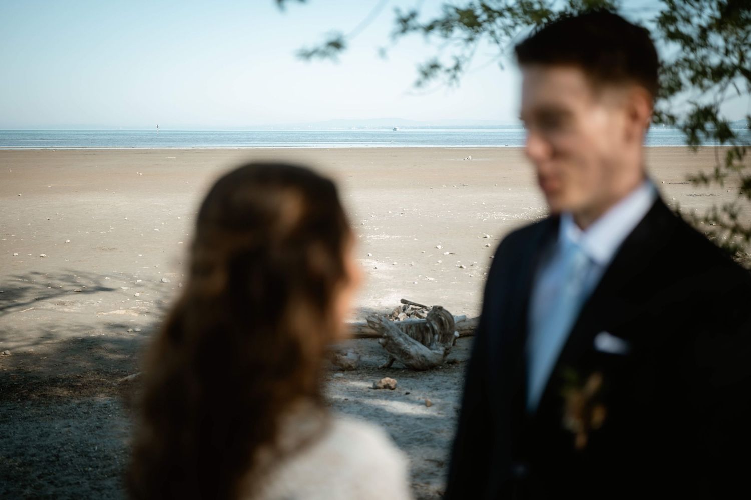 Unscharfes Brautpaar mit Blick auf den Bodensee und Treibholz im Vordergrund – Hochzeit Jacqueline & Clemens