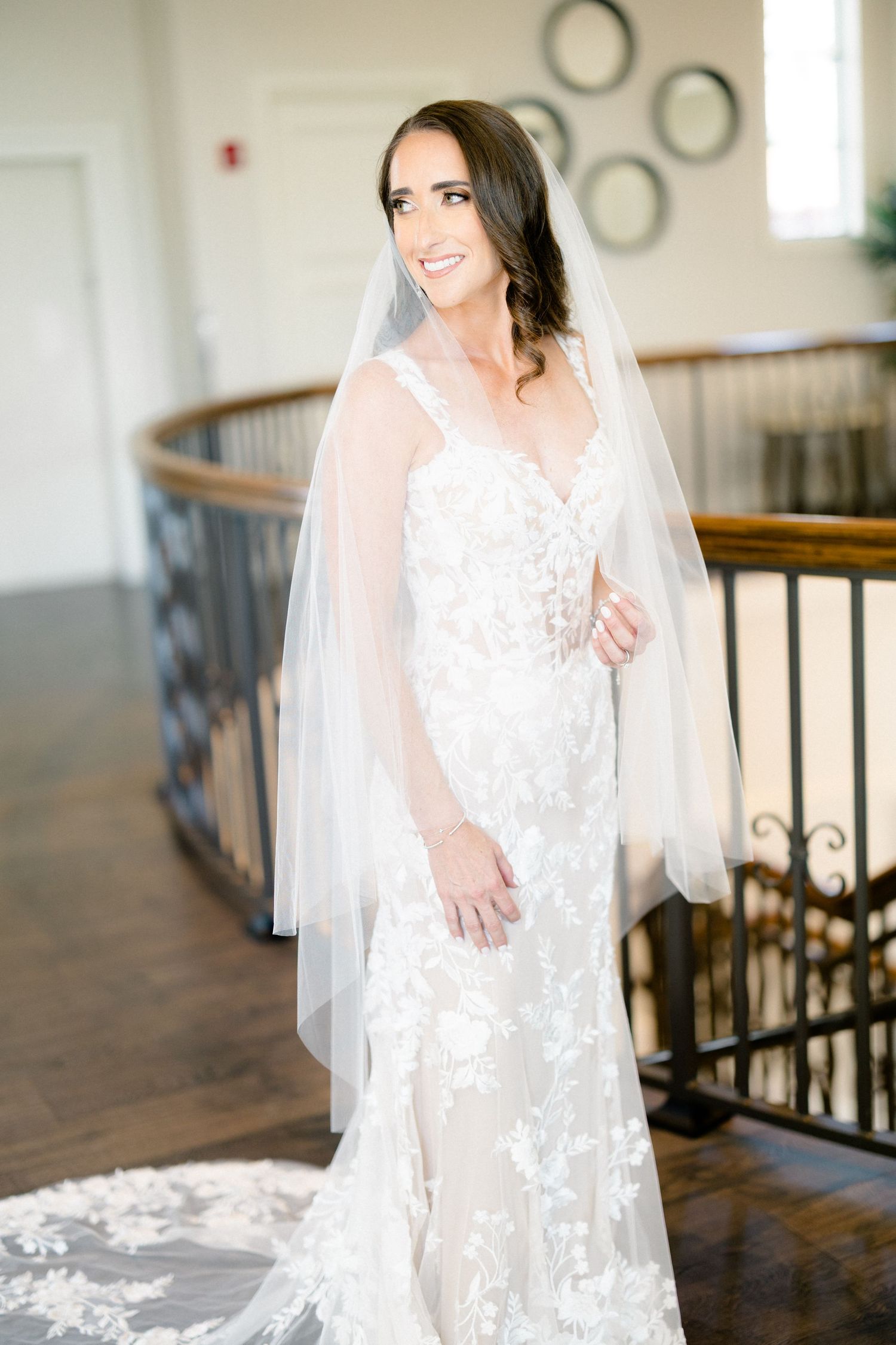 Bride in lace wedding gown and veil smiling on elegant staircase.