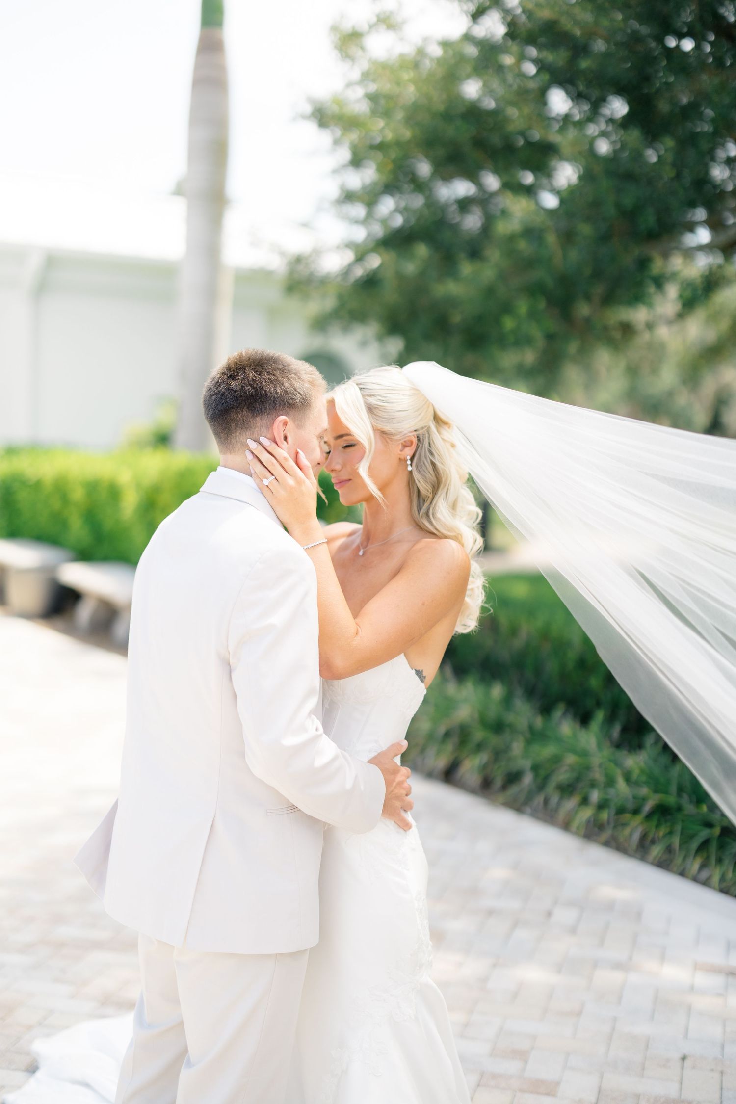 Bride and groom share romantic moment, her veil flowing in breeze, during outdoor wedding portrait session.