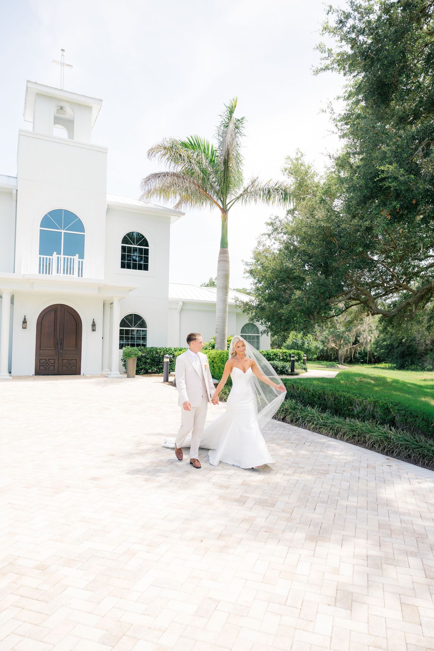 Bride and groom hold hands walking outside a white chapel with palm trees on a bright sunny wedding day.