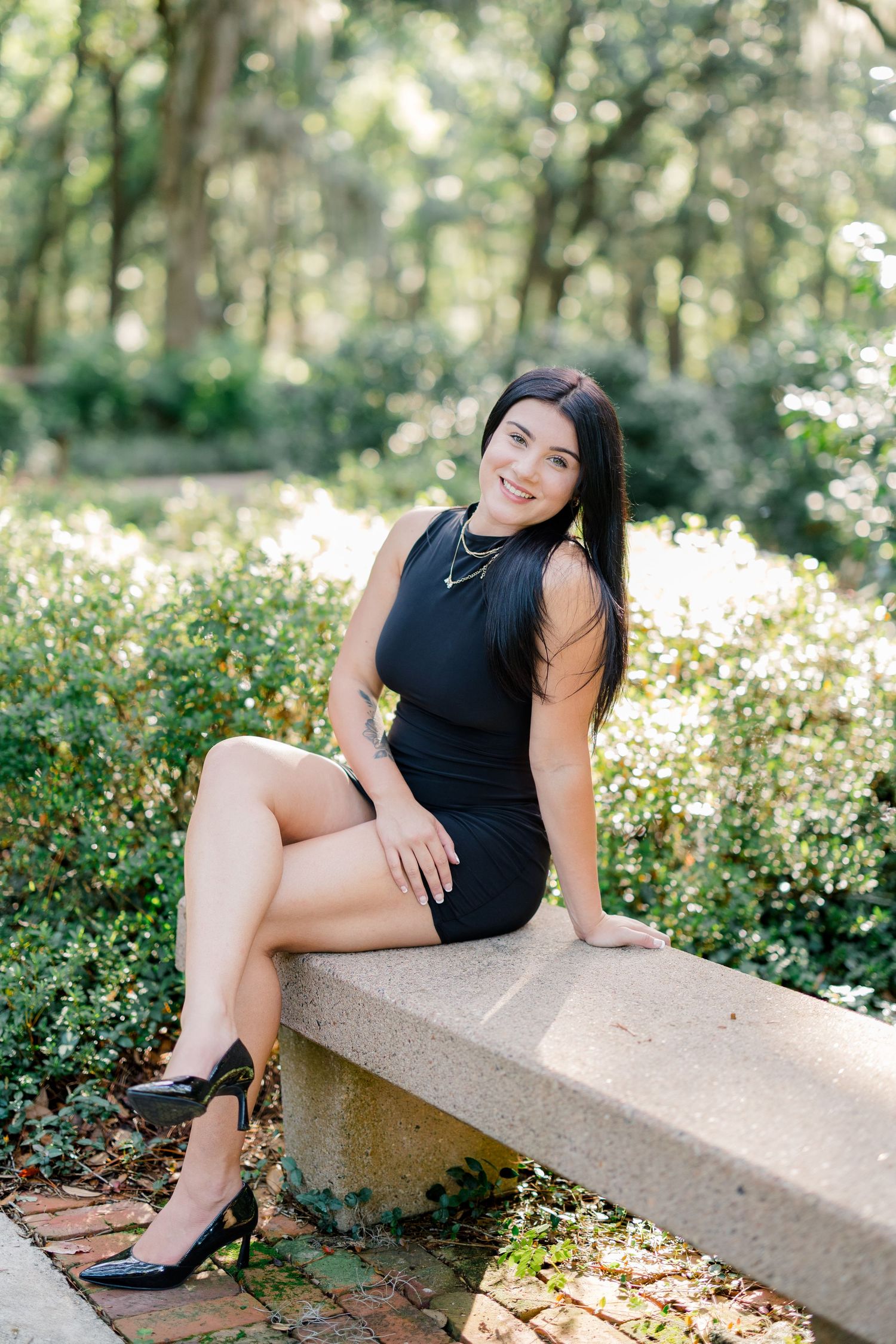 Young woman in black mini dress and heels sits on stone bench smiling in a lush green park setting.