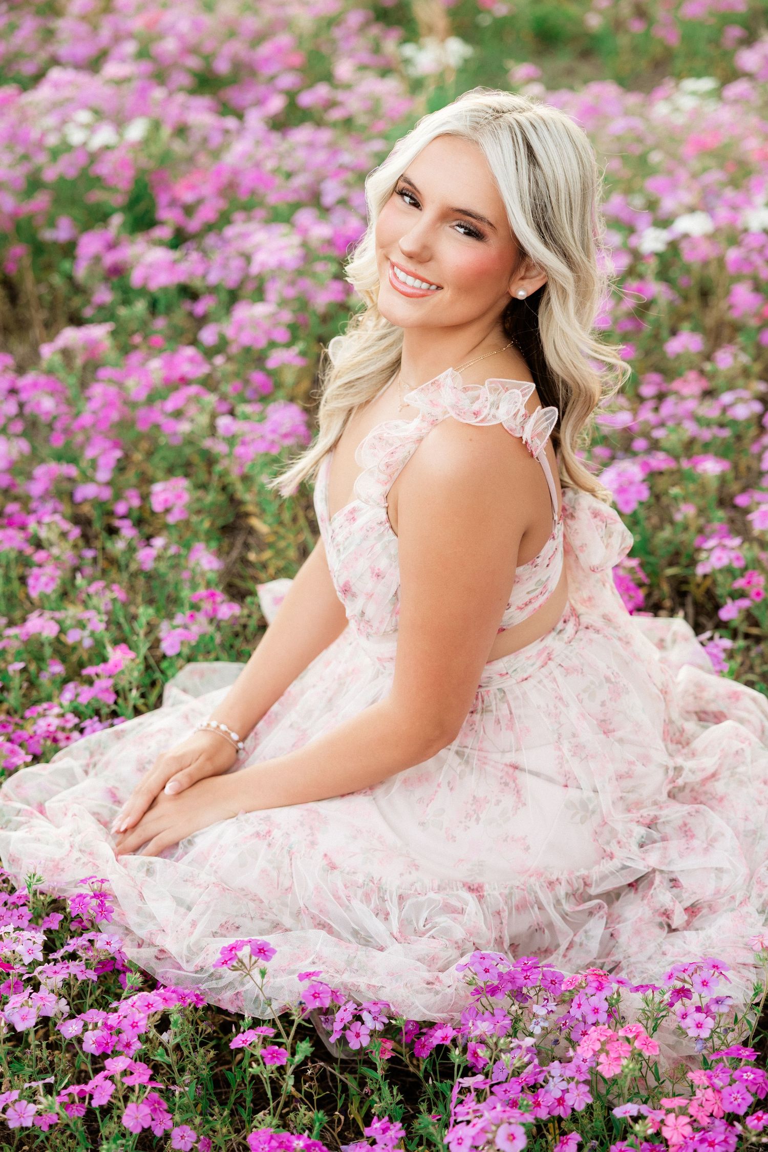 Smiling blonde woman in a pink floral dress sitting in a field of pink flowers.