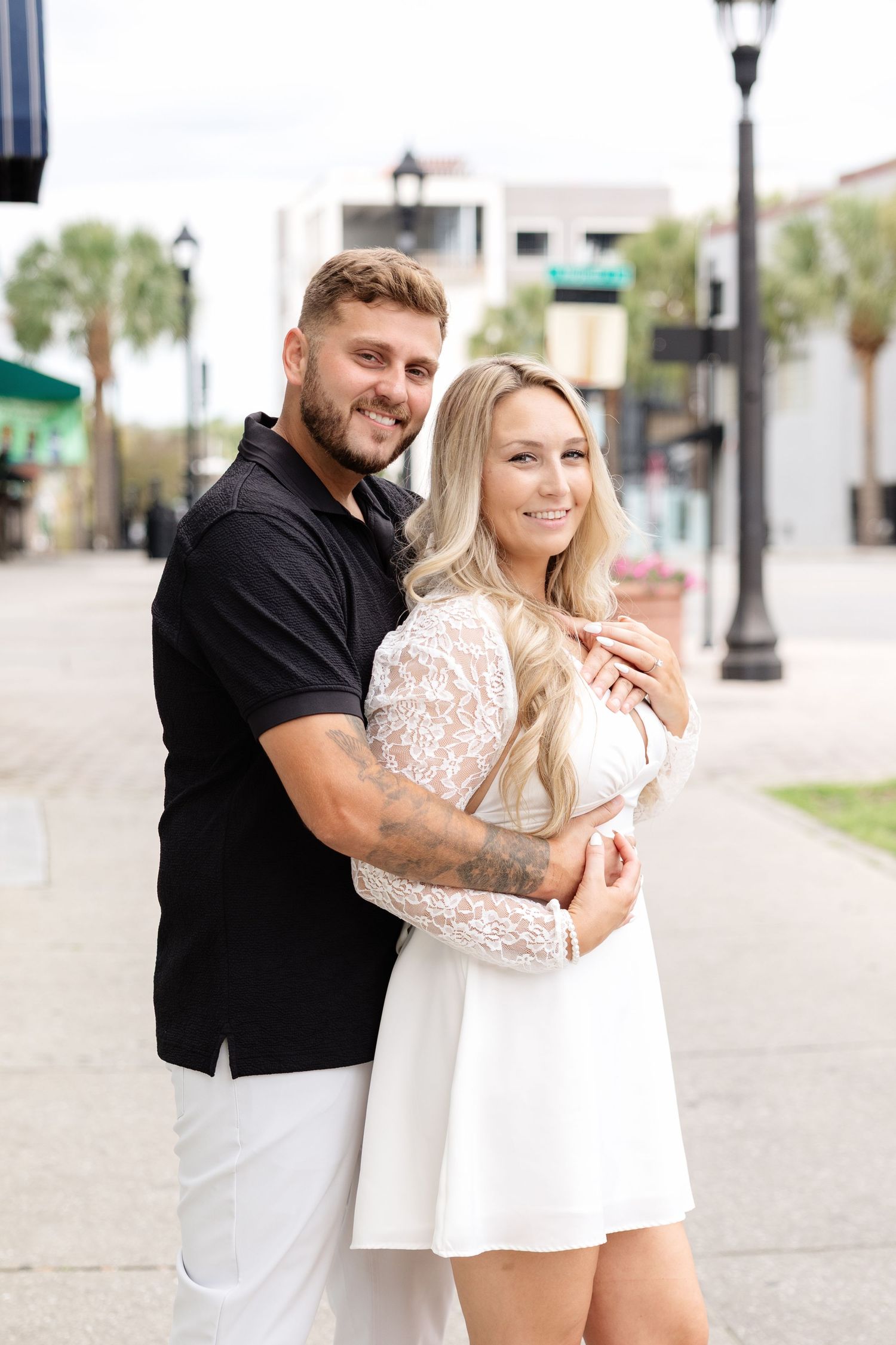 A smiling couple embraces on a sunny urban sidewalk, the man in black and the woman in a white lace dress.