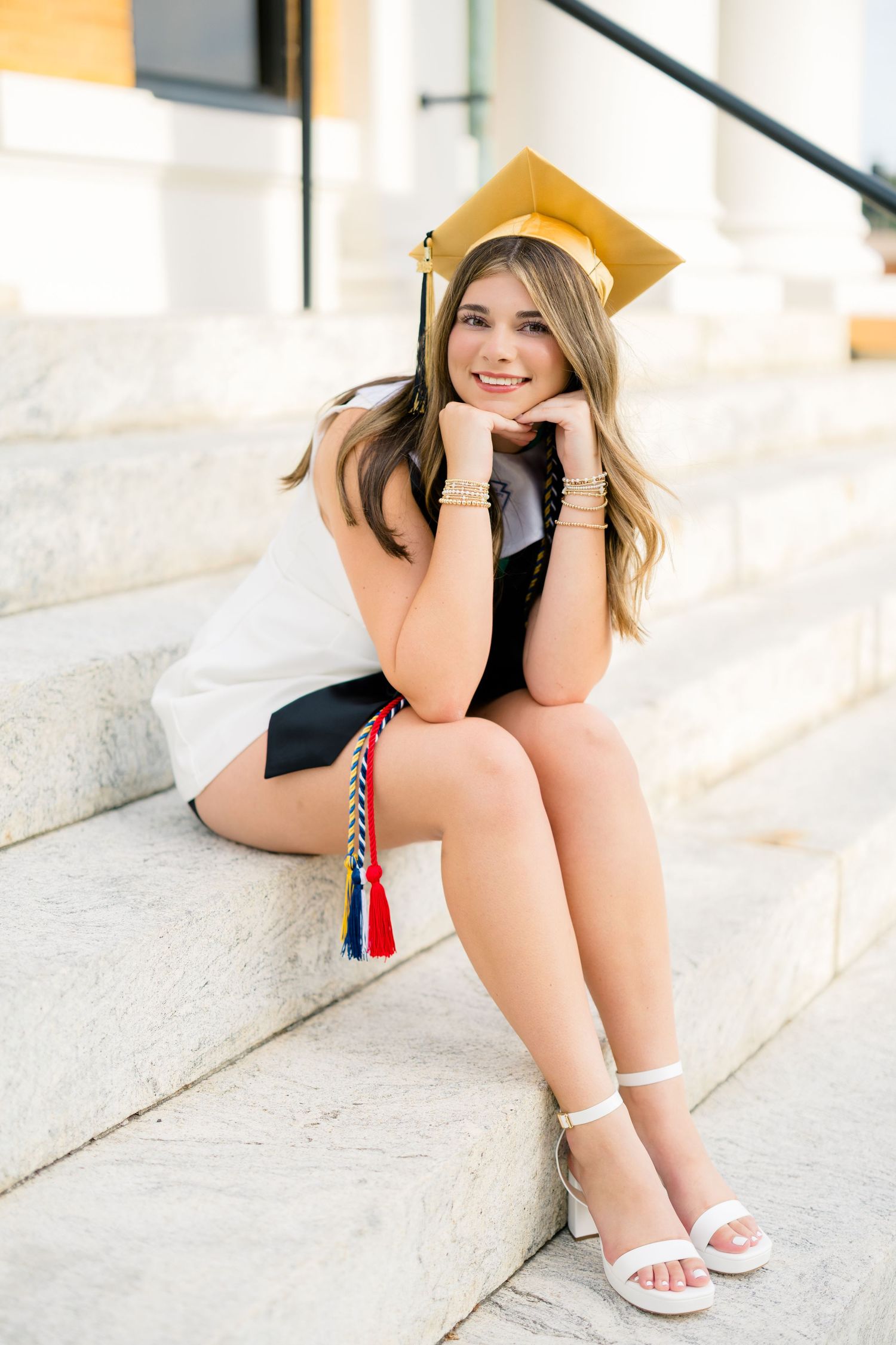 Smiling graduate girl sitting on marble steps wearing gold cap, white dress, and colorful honor cords.