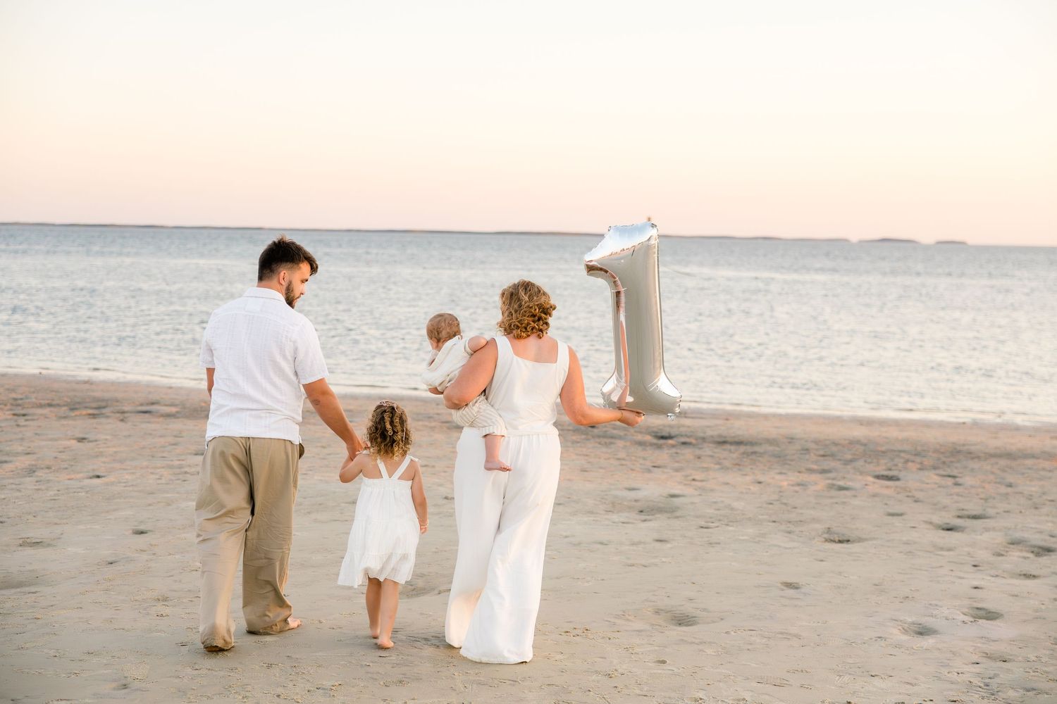 Family of four walking on sandy beach at sunset, woman holding number 1 balloon, dressed in white and beige outfits.
