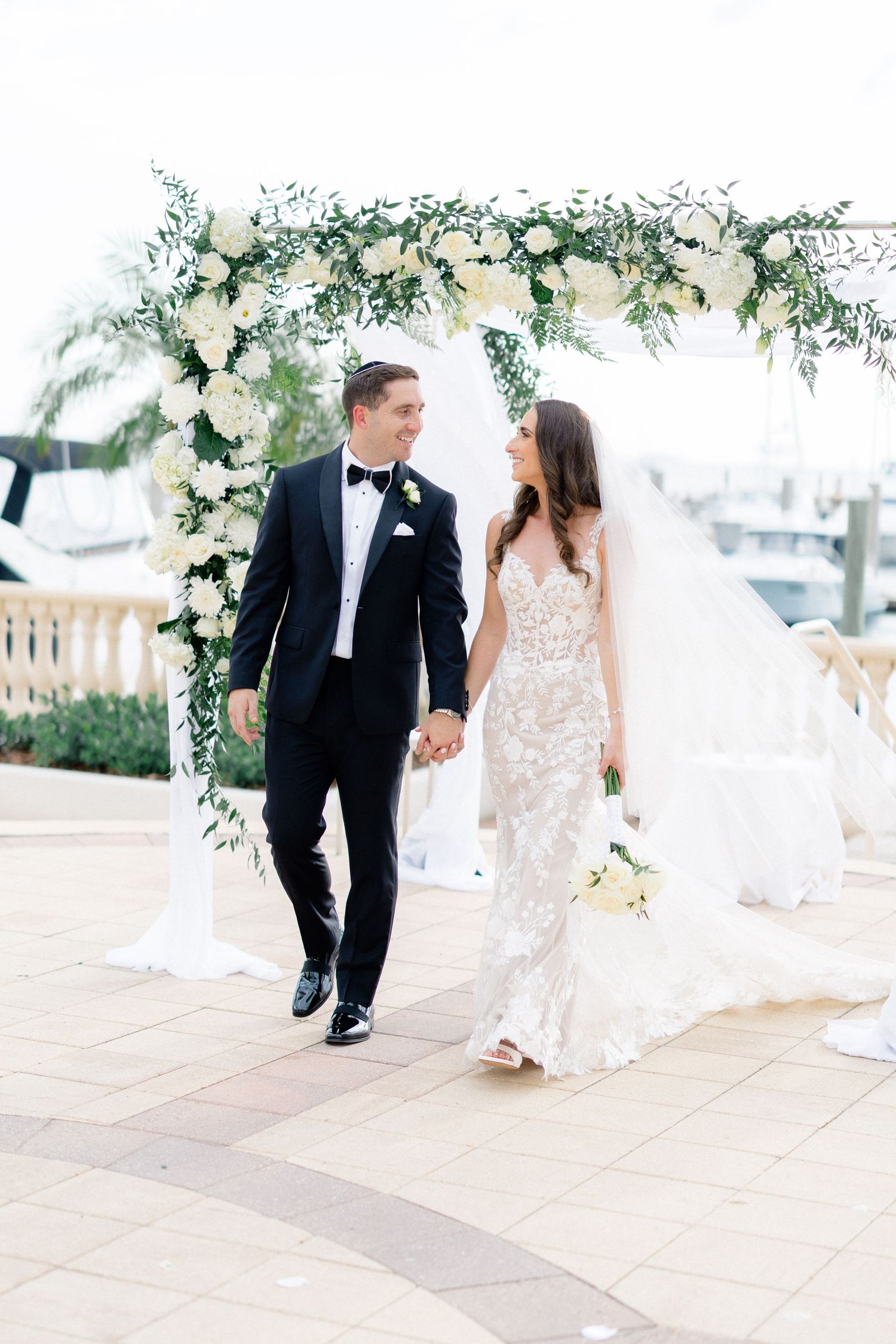 Bride and groom walk hand-in-hand under a floral arch with white roses and greenery at a waterfront wedding venue.