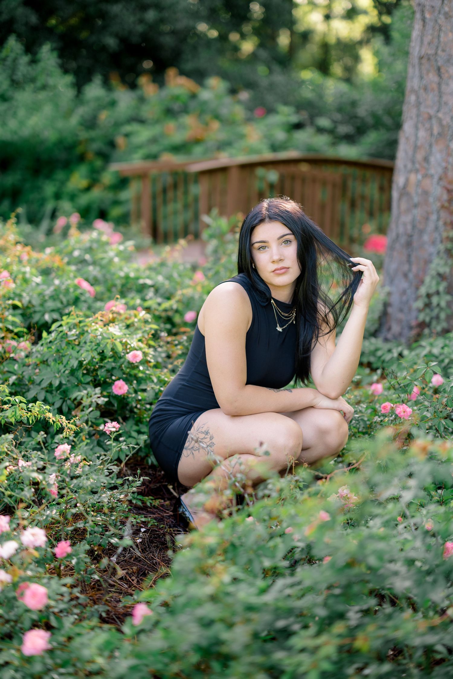 Young woman in black dress crouching among pink roses in a lush garden with a wooden bridge backdrop.