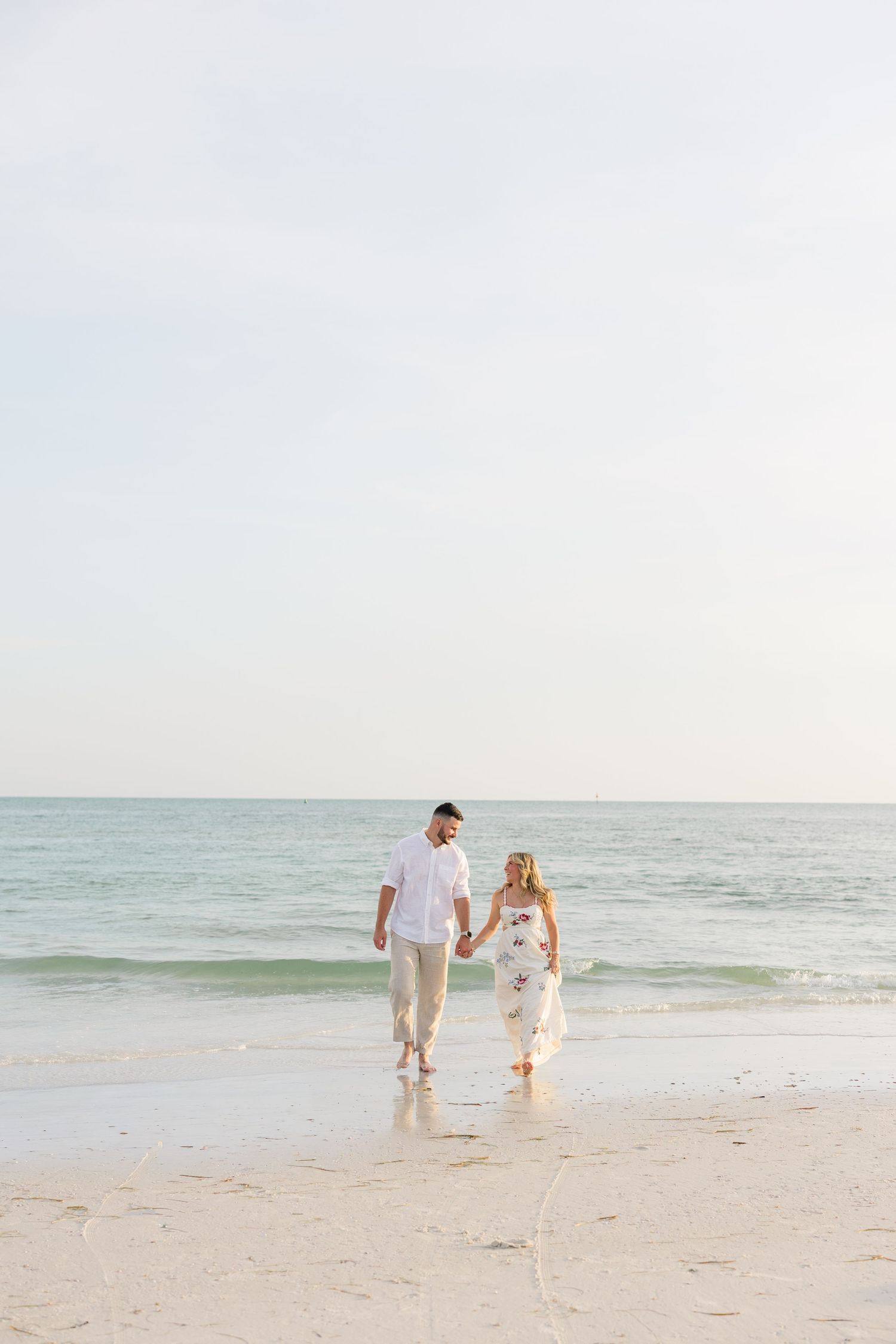 Couple holding hands walking on a sandy beach near the ocean shore, wearing white outfits on a calm, bright day.