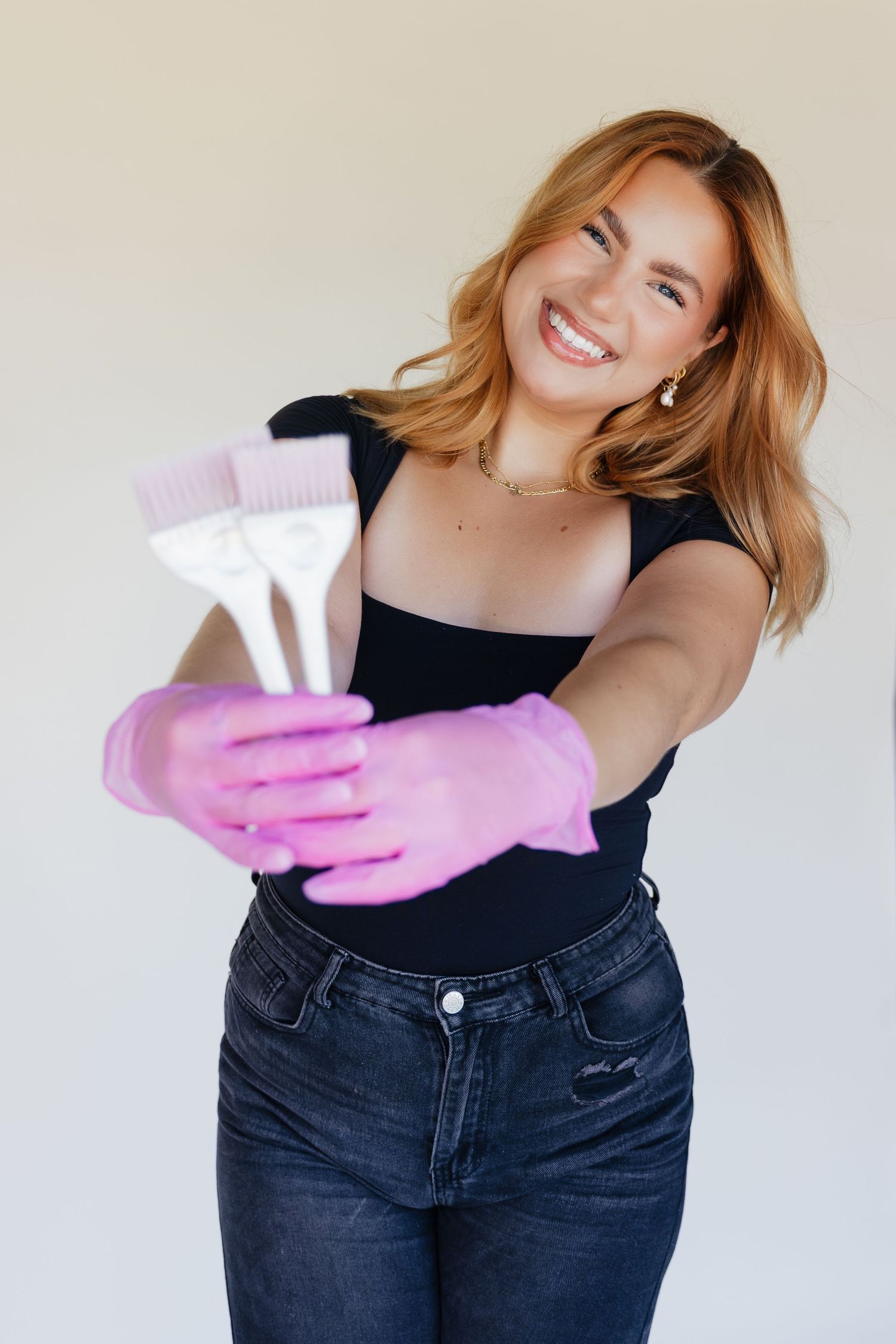 Smiling woman in black top and jeans wearing pink gloves, holding hair dye brushes toward camera on white background.