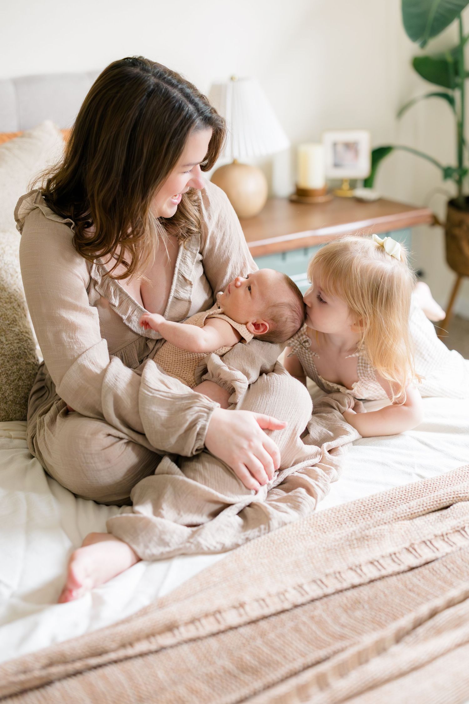 A mother sits on a bed holding a newborn while a young blonde toddler girl leans in to give the baby a kiss.
