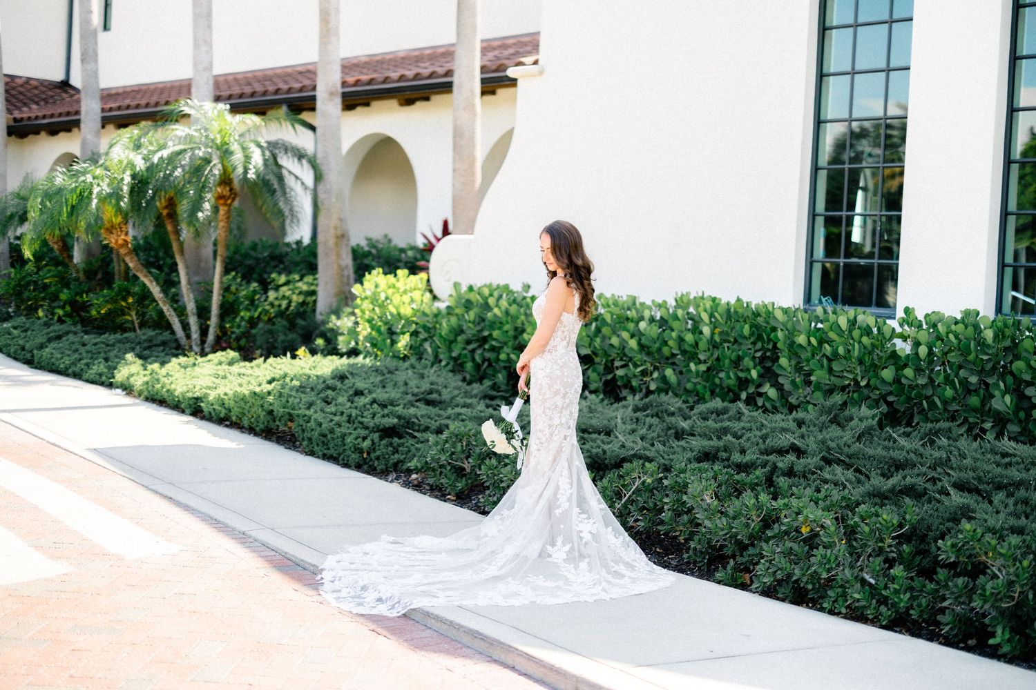Bride in lace gown with long train walks along path beside manicured hedges at venue.
