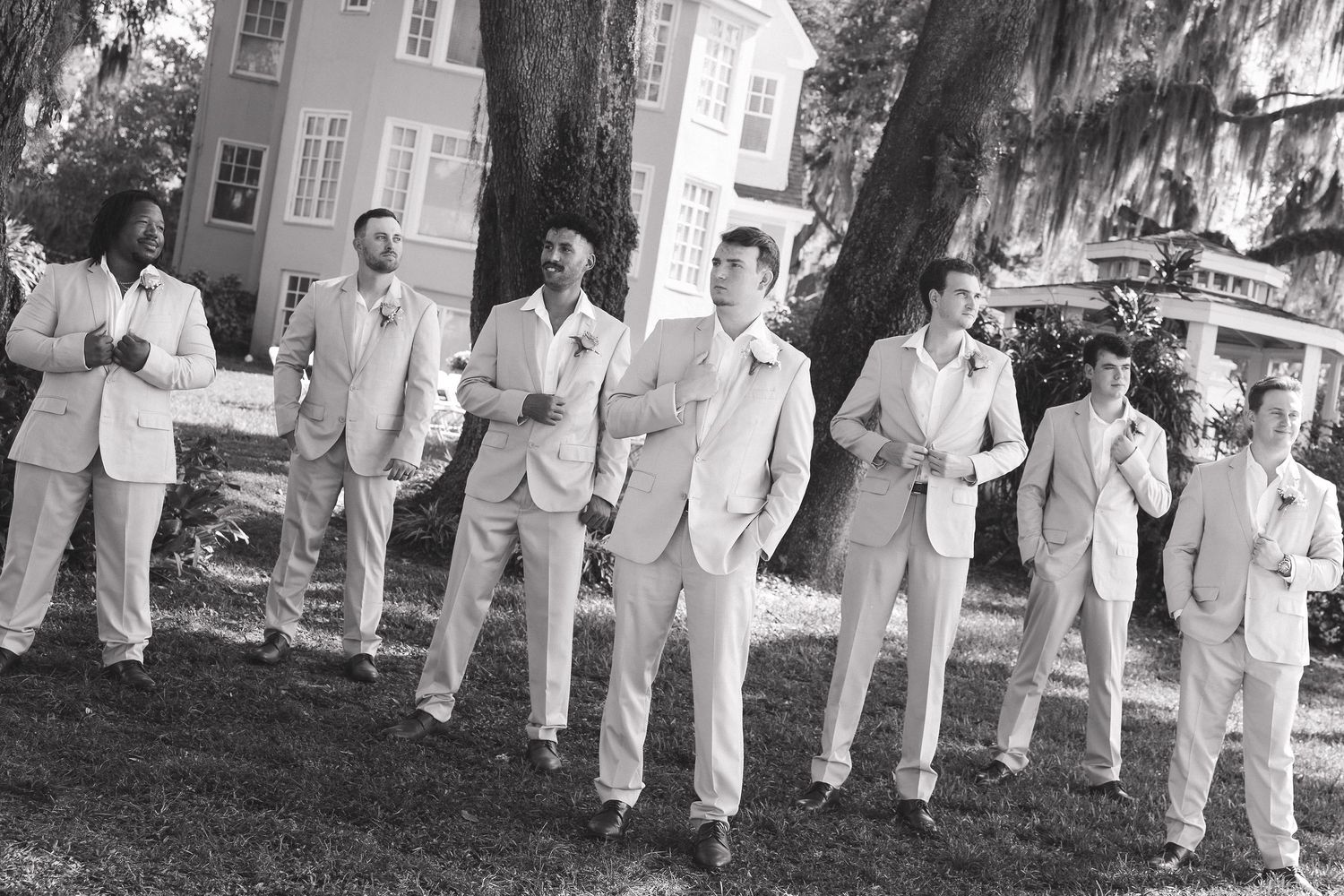 Wedding party members in light suits stand in formation on grass near a building.