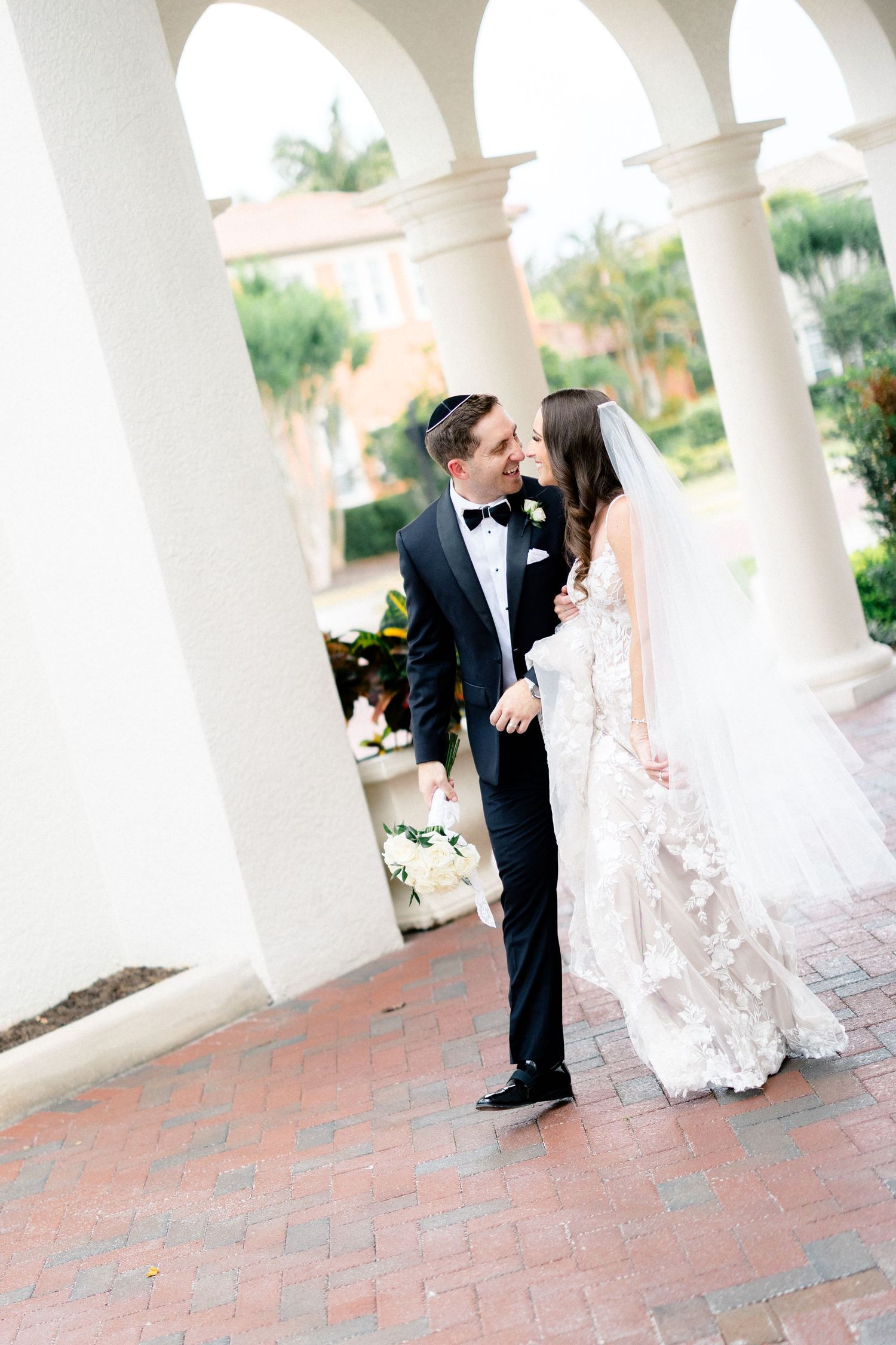 A bride and groom share a romantic moment under elegant white columns at an outdoor wedding venue.