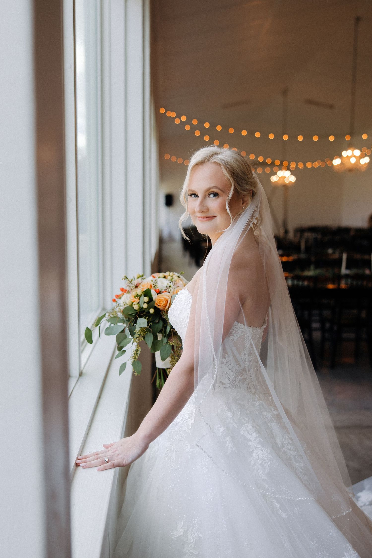 Elegant bride in lace gown and veil poses by window with string lights in background.