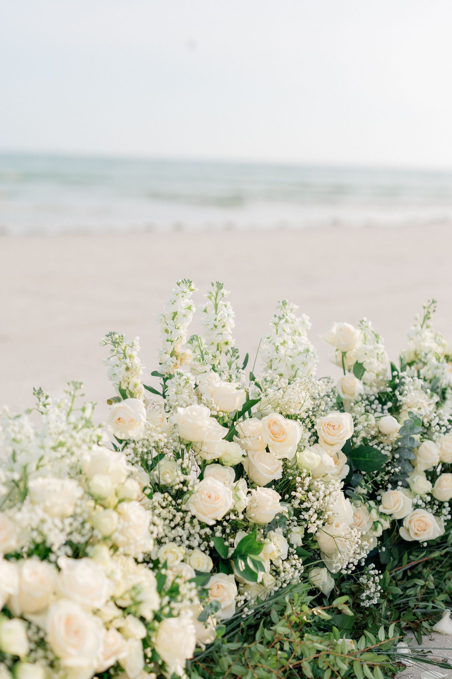 White roses and baby's breath floral arrangement on a sandy beach with ocean waves in the background.