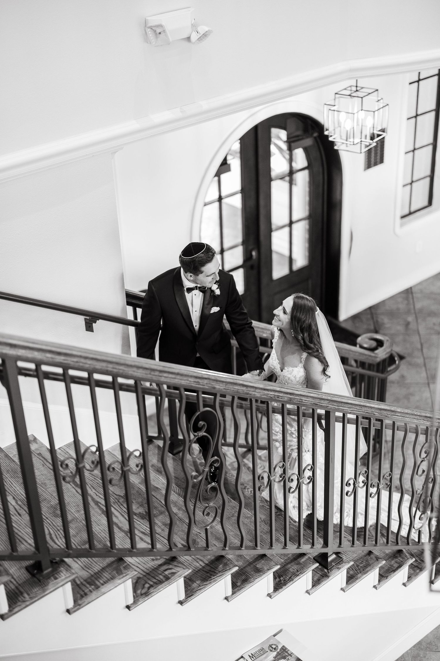Black and white photo of a bride and groom sharing a romantic moment on an elegant staircase with ornate iron railings.