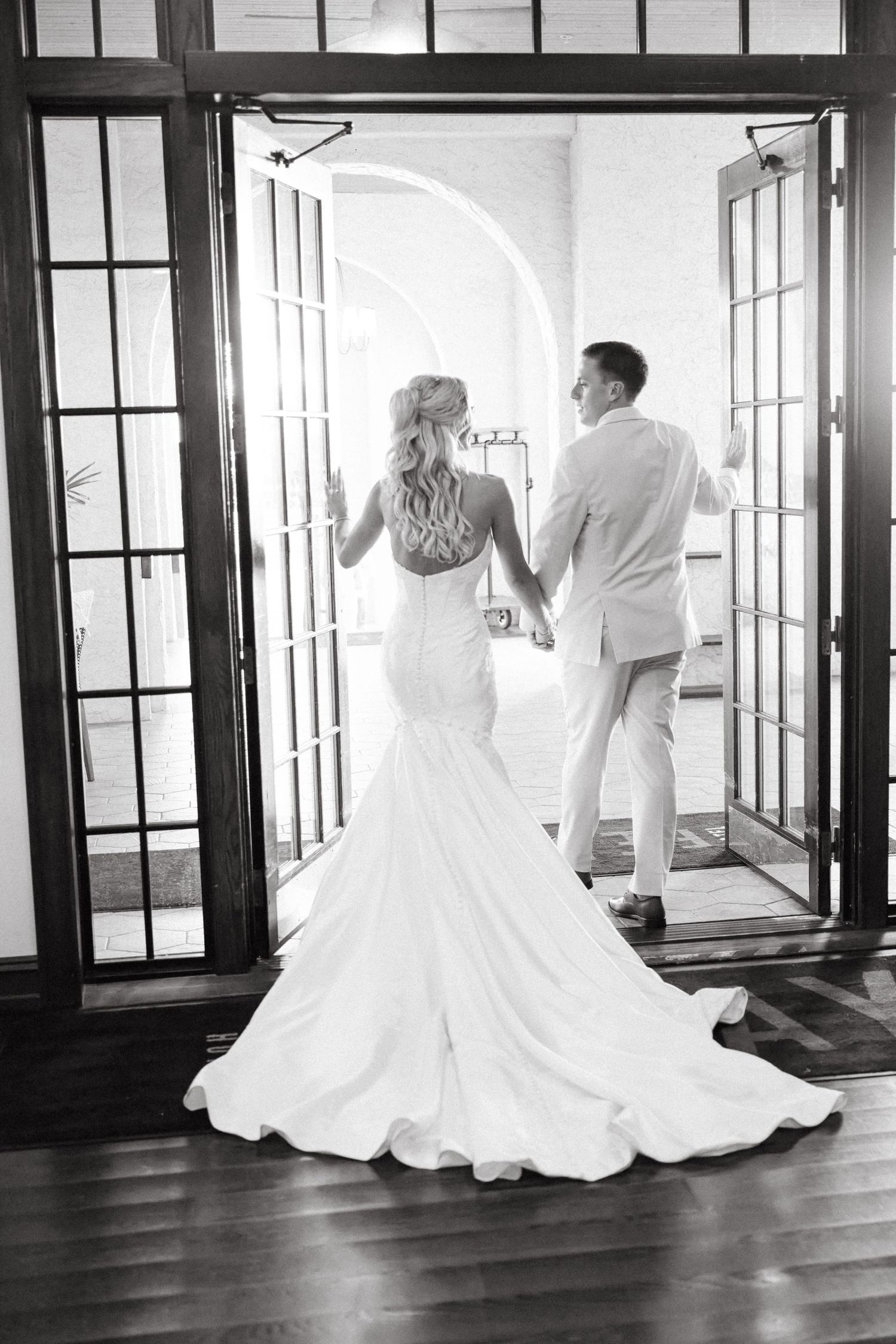 Black and white photo of bride in mermaid gown and groom holding hands walking through elegant French doors.