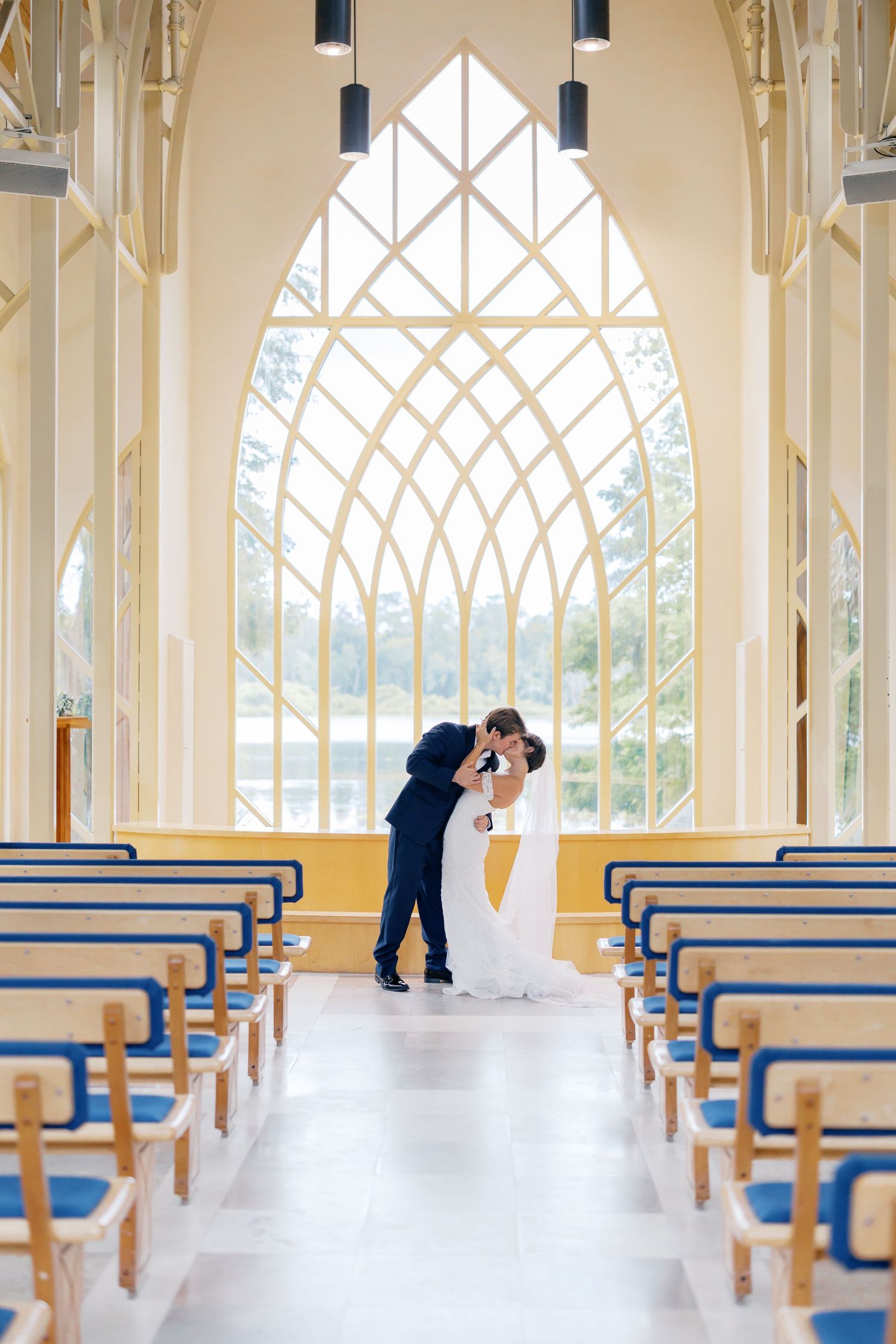 Bride and groom share a romantic kiss at the altar of an elegant chapel with gothic arched windows and blue pew seating.