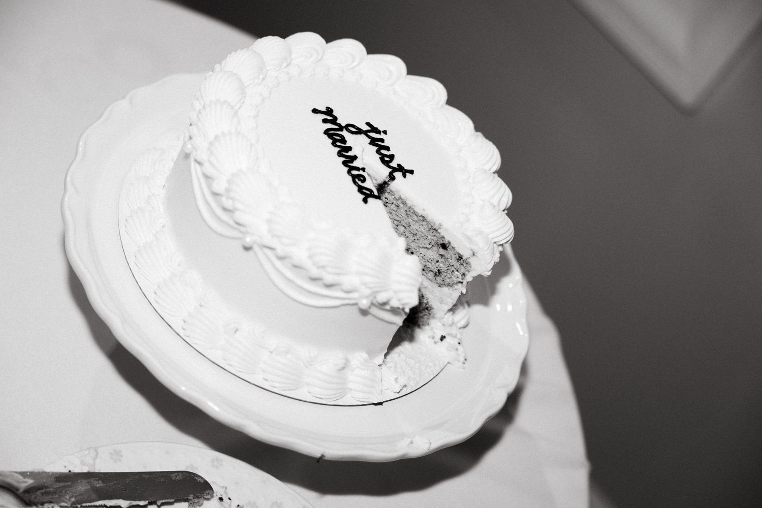 Black and white photo of a round white wedding cake with cursive writing, partially sliced, on a decorative cake stand.