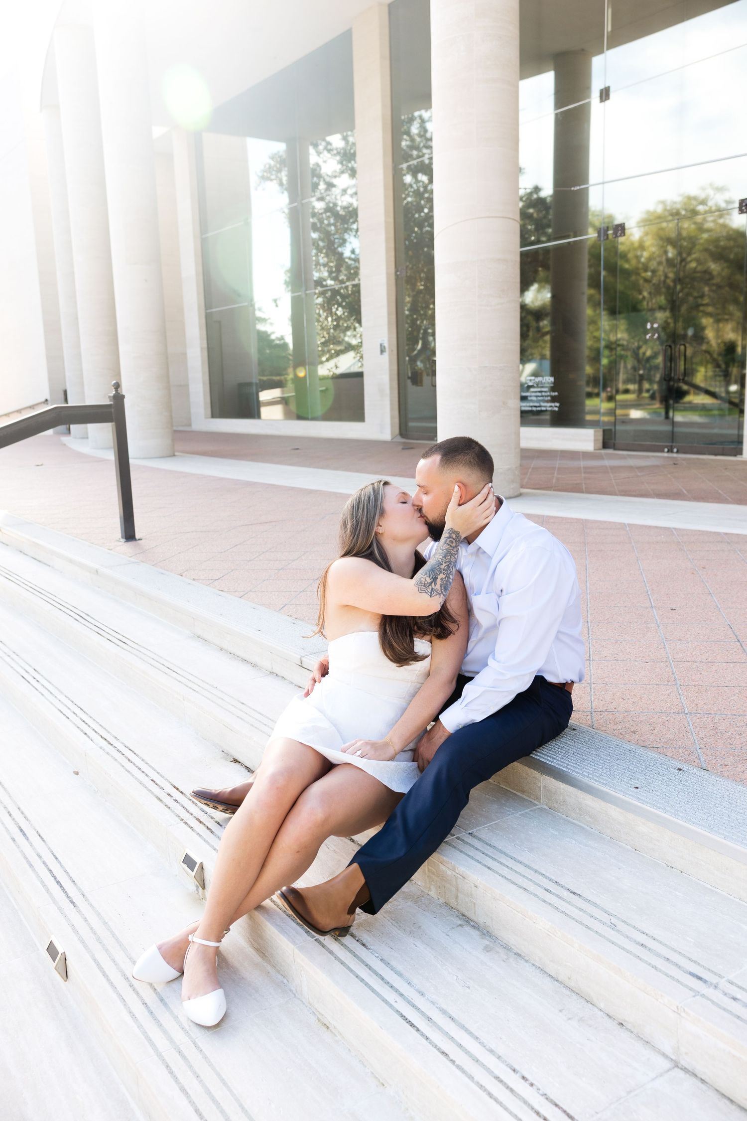 Couple sharing a romantic kiss while sitting on marble steps outside a modern glass building in bright sunlight.