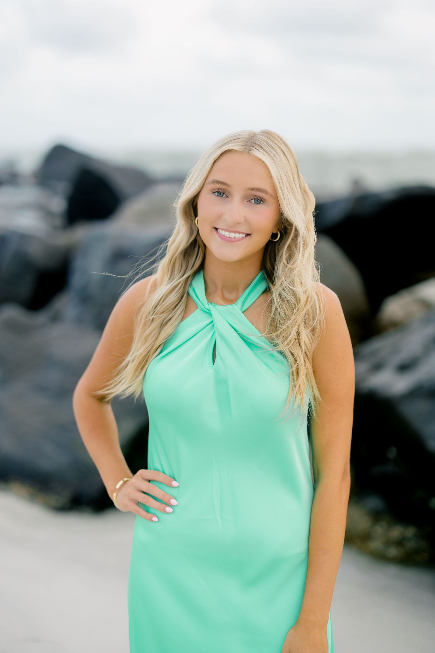 Young blonde woman in mint green halter dress smiling on beach with rocks in background.