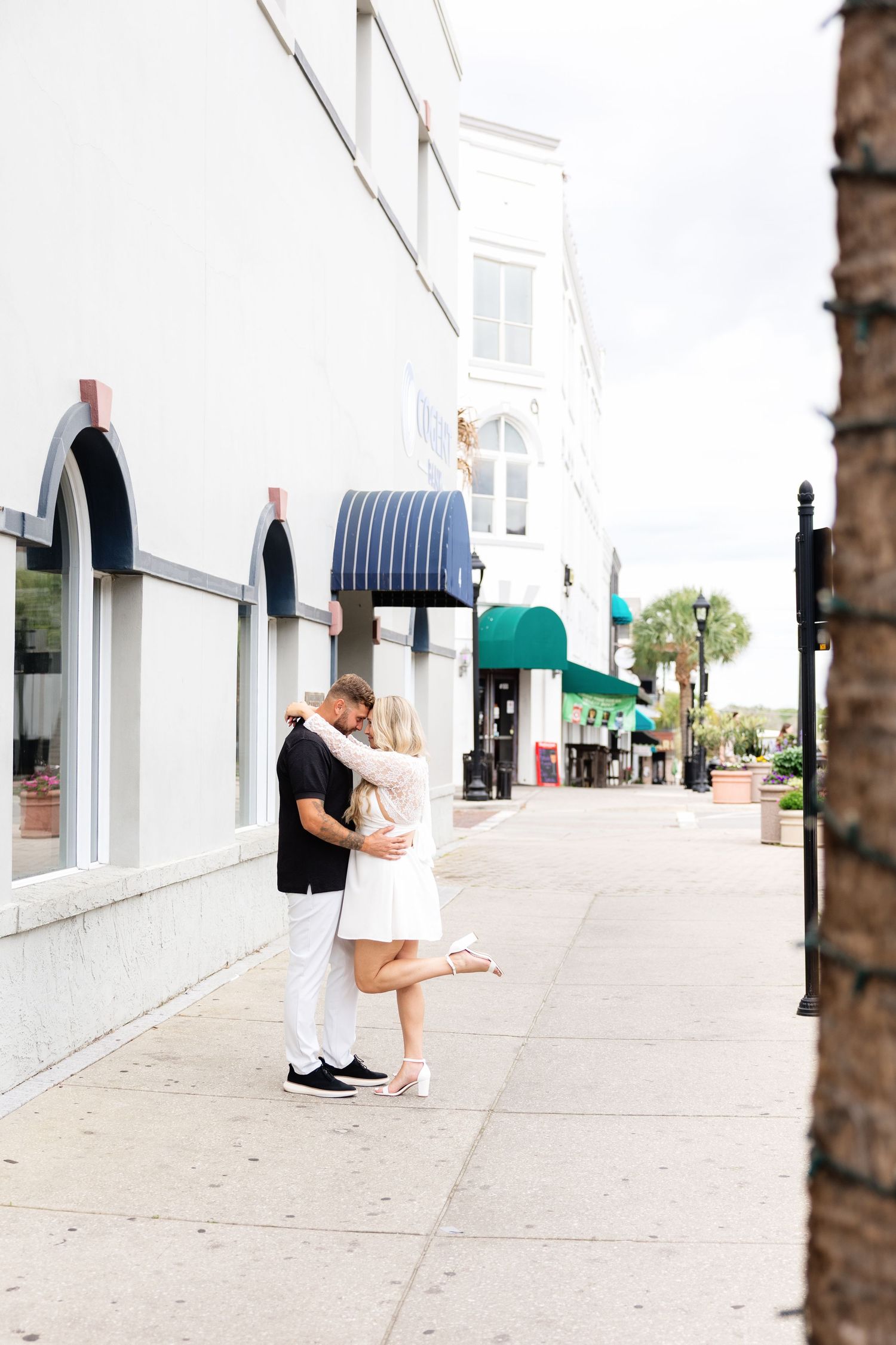 Couple sharing a romantic kiss on a sunny downtown sidewalk, woman in white skirt lifting her foot, beside a white building.