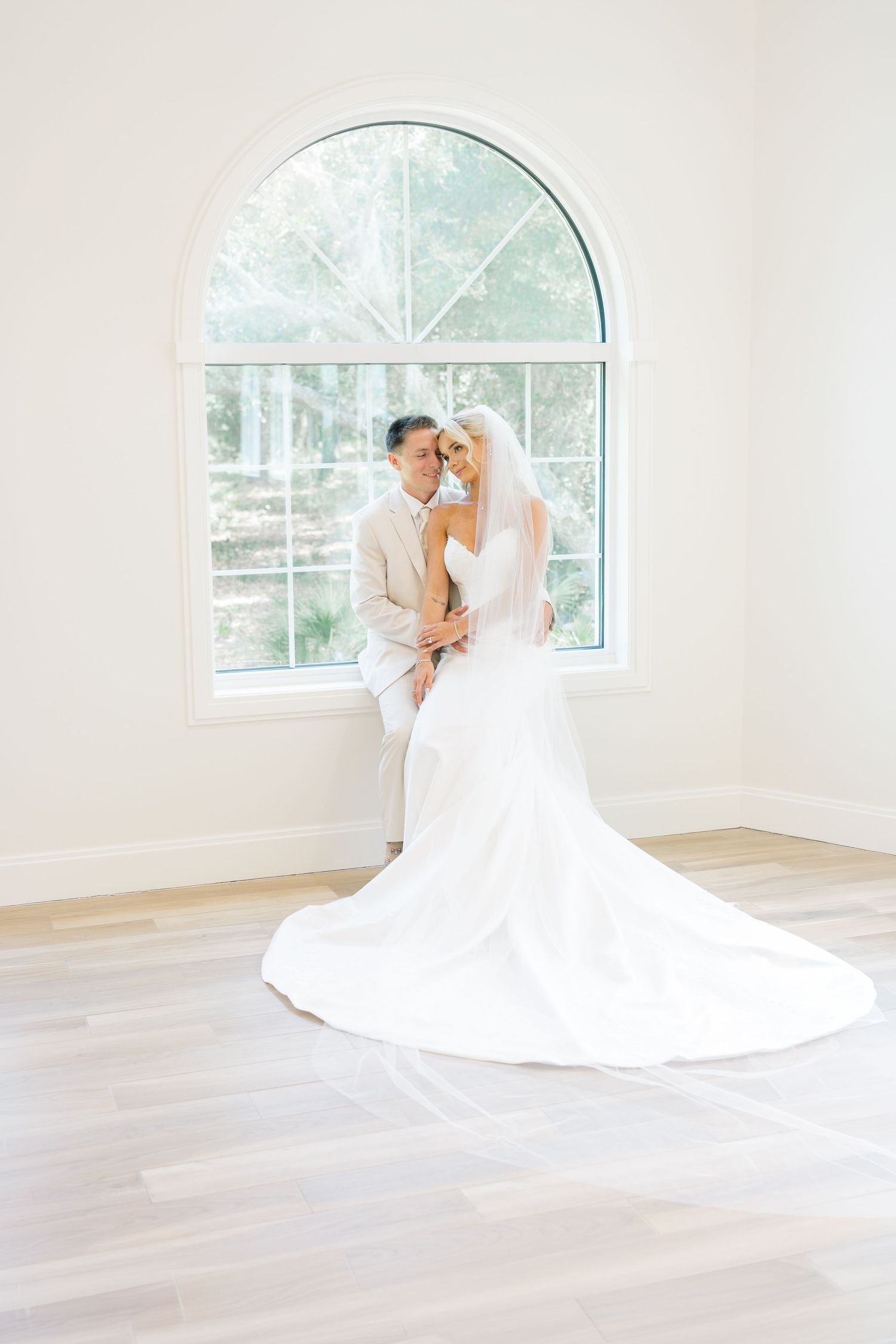 Bride and groom embrace before arched window in bright white room, bride in elegant white gown with long train.
