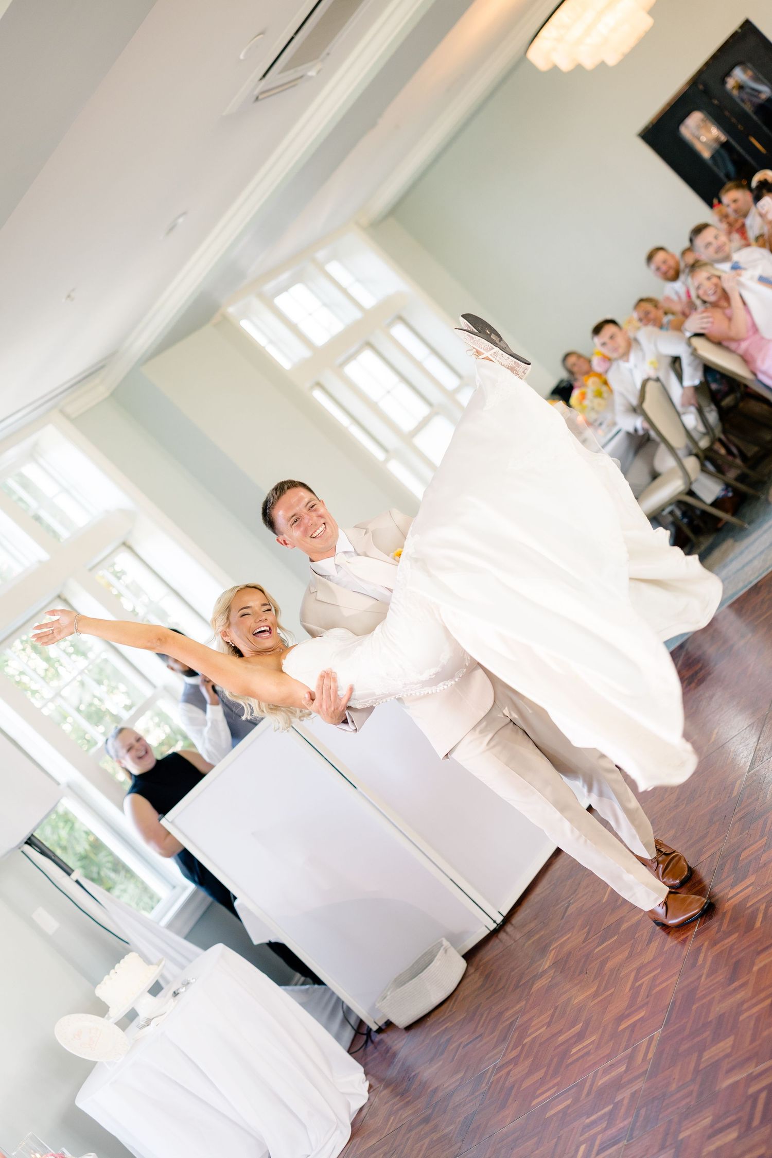 Bride and groom share joyful first dance at wedding reception with guests watching and smiling in background.
