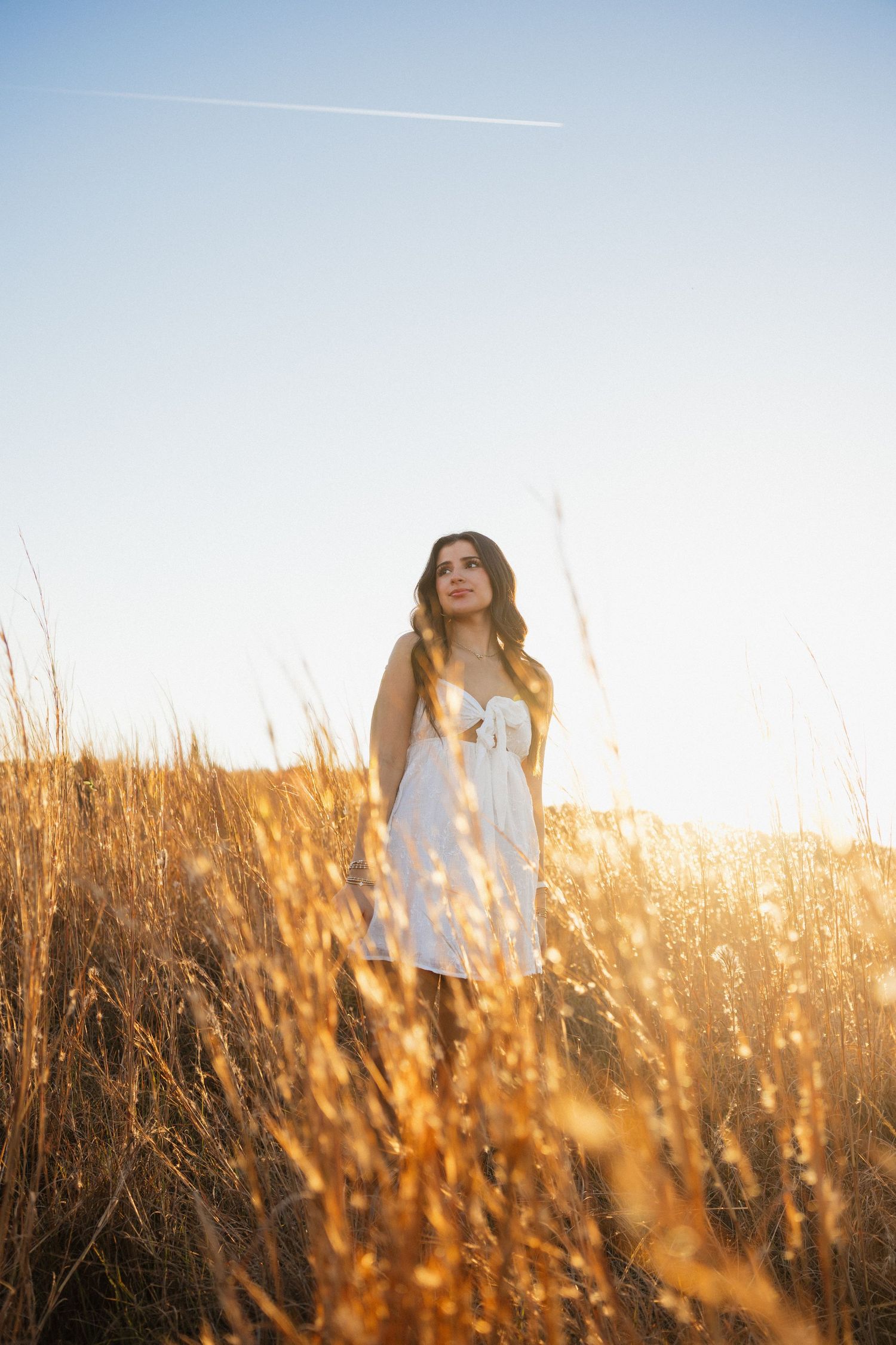 Young woman in white dress standing in golden wheat field at sunset, looking up at clear blue sky.