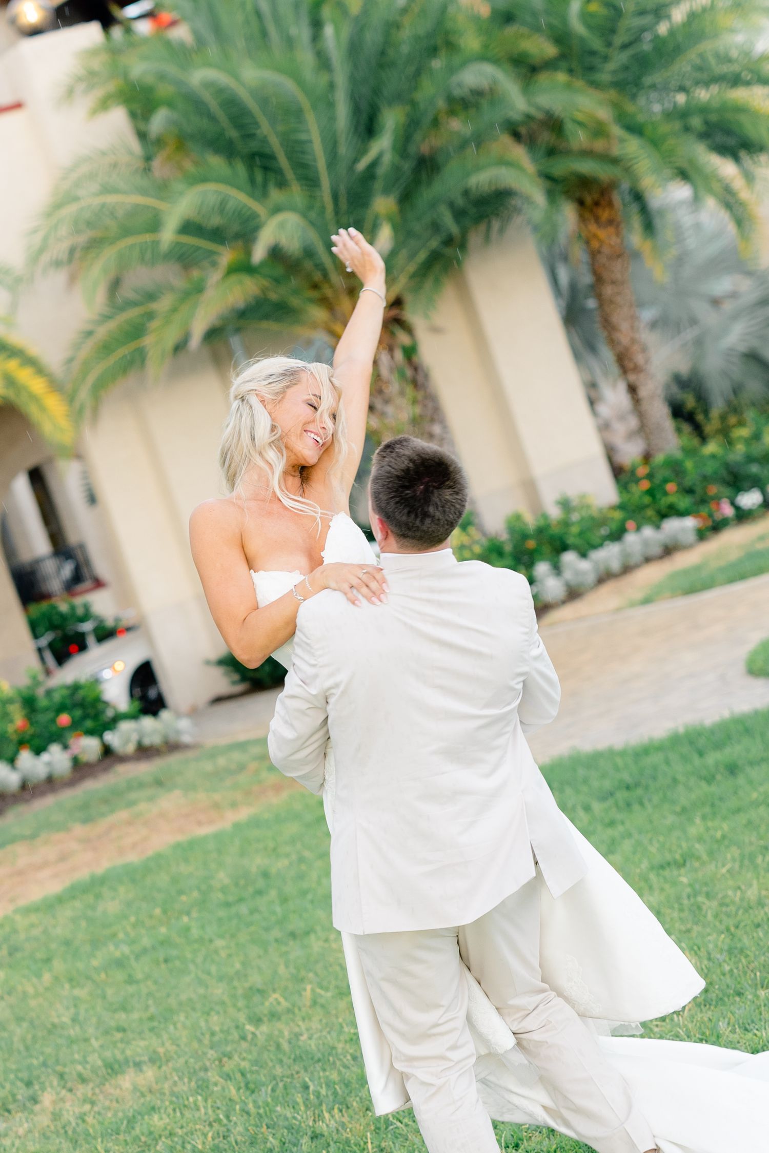 Groom in white suit lifting bride in strapless gown outdoors near palm trees during wedding celebration.