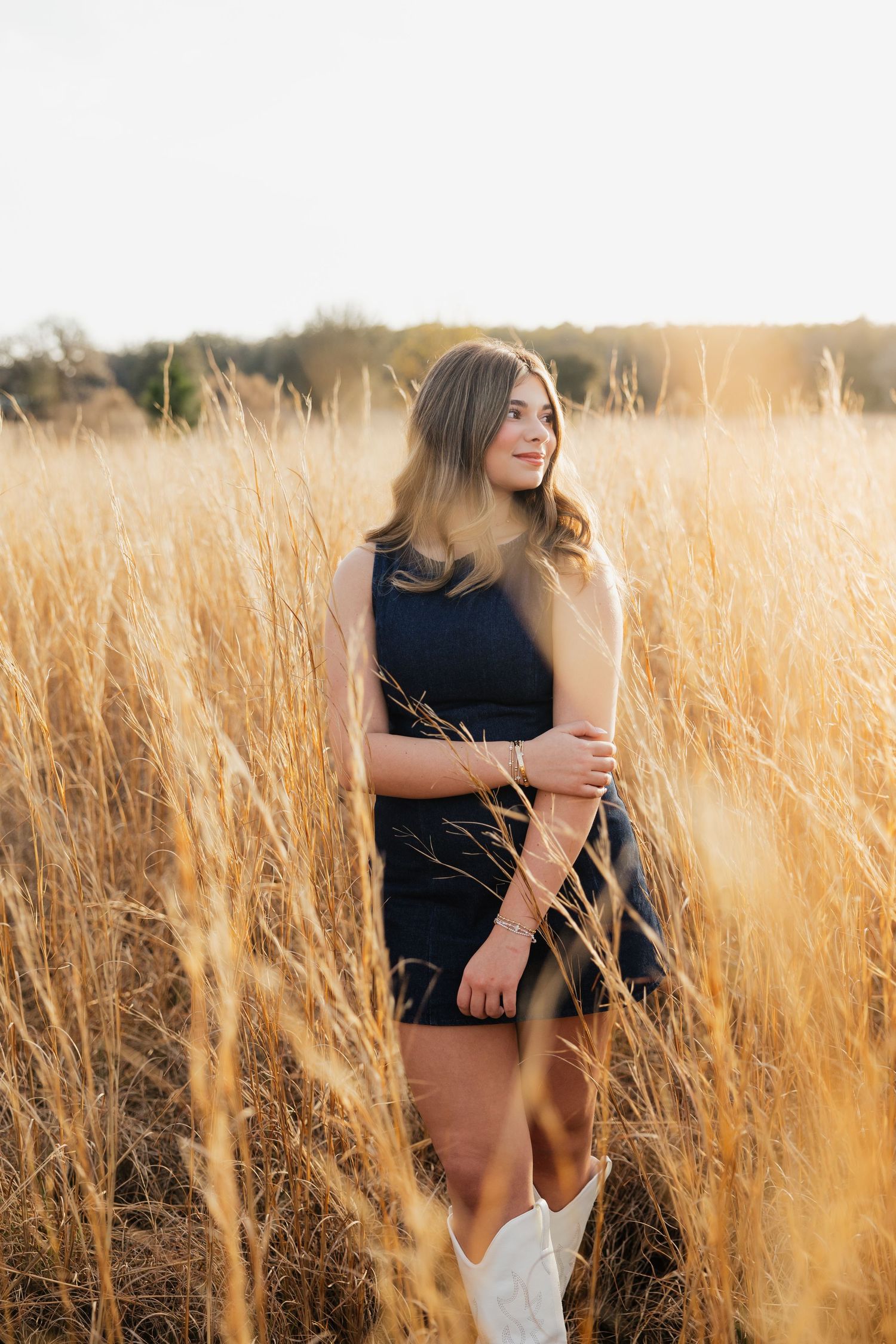 Young woman in navy dress and white cowboy boots standing in golden field at sunset.