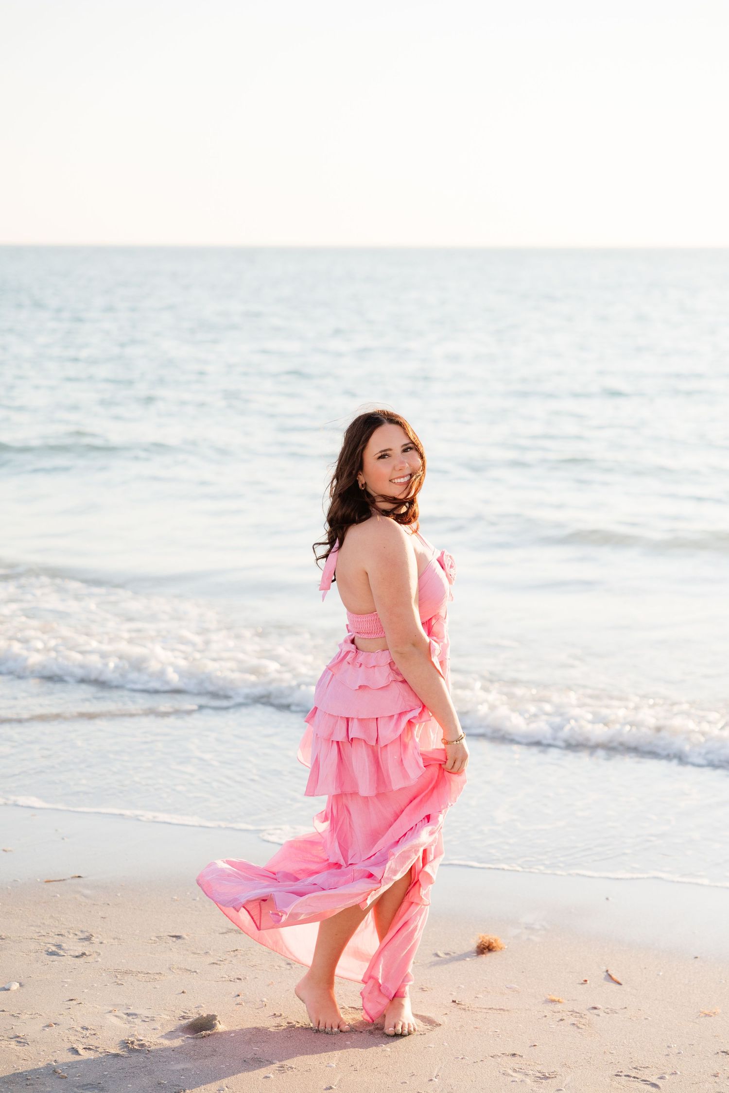Woman in pink ruffled dress walking barefoot on sandy beach with ocean waves behind her.