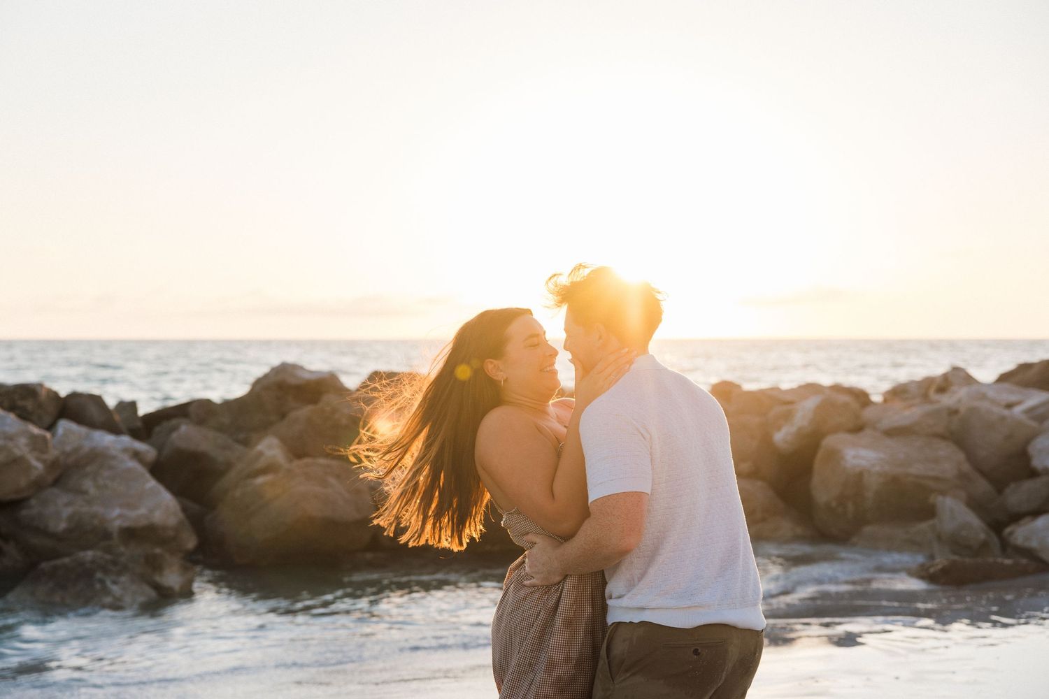 Couple sharing a romantic moment at sunset by rocky beach, golden light glowing behind them as they embrace near the ocean.