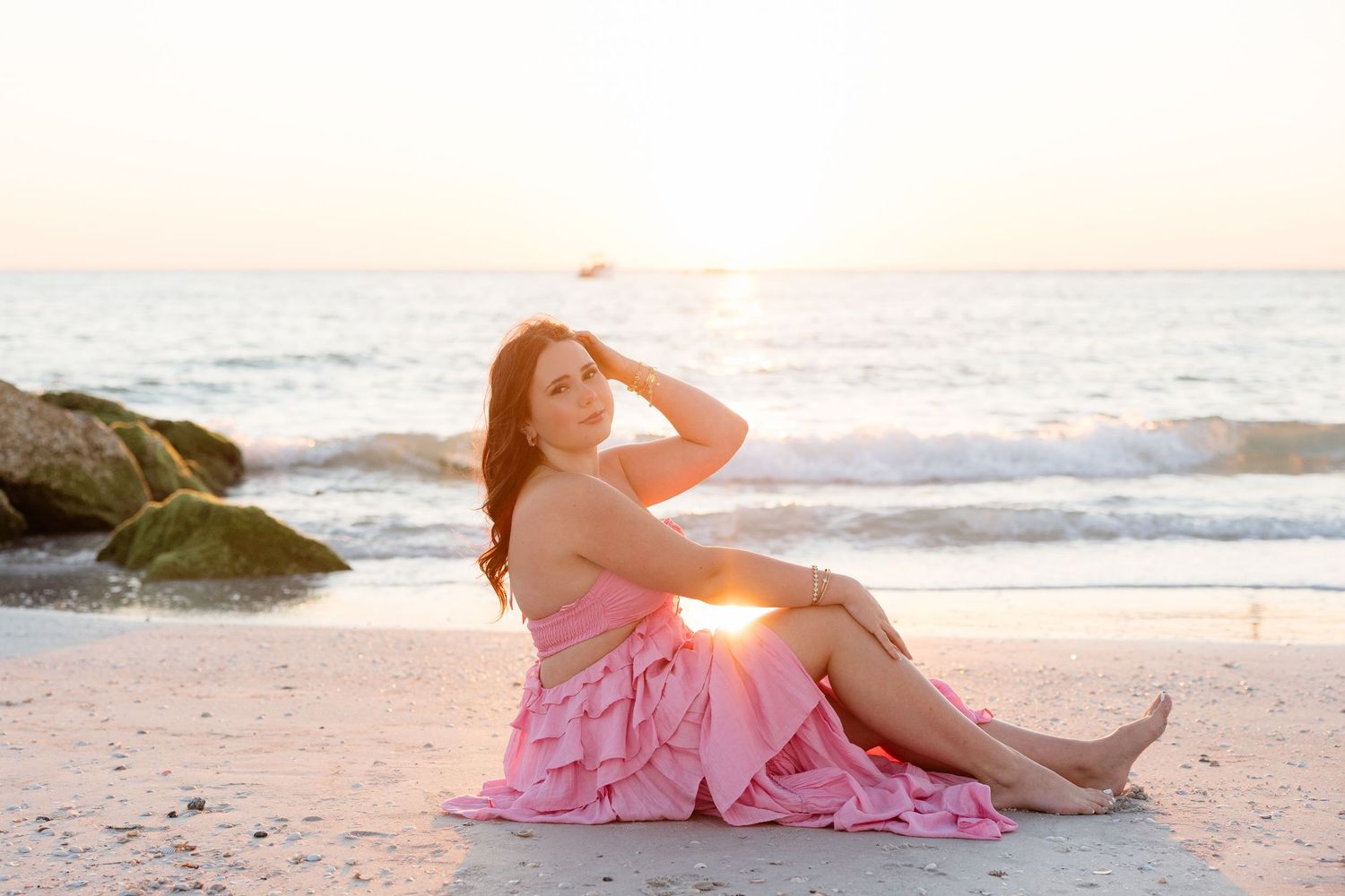 Woman in pink ruffled dress sitting on sandy beach at sunset with ocean waves and rocks in background.