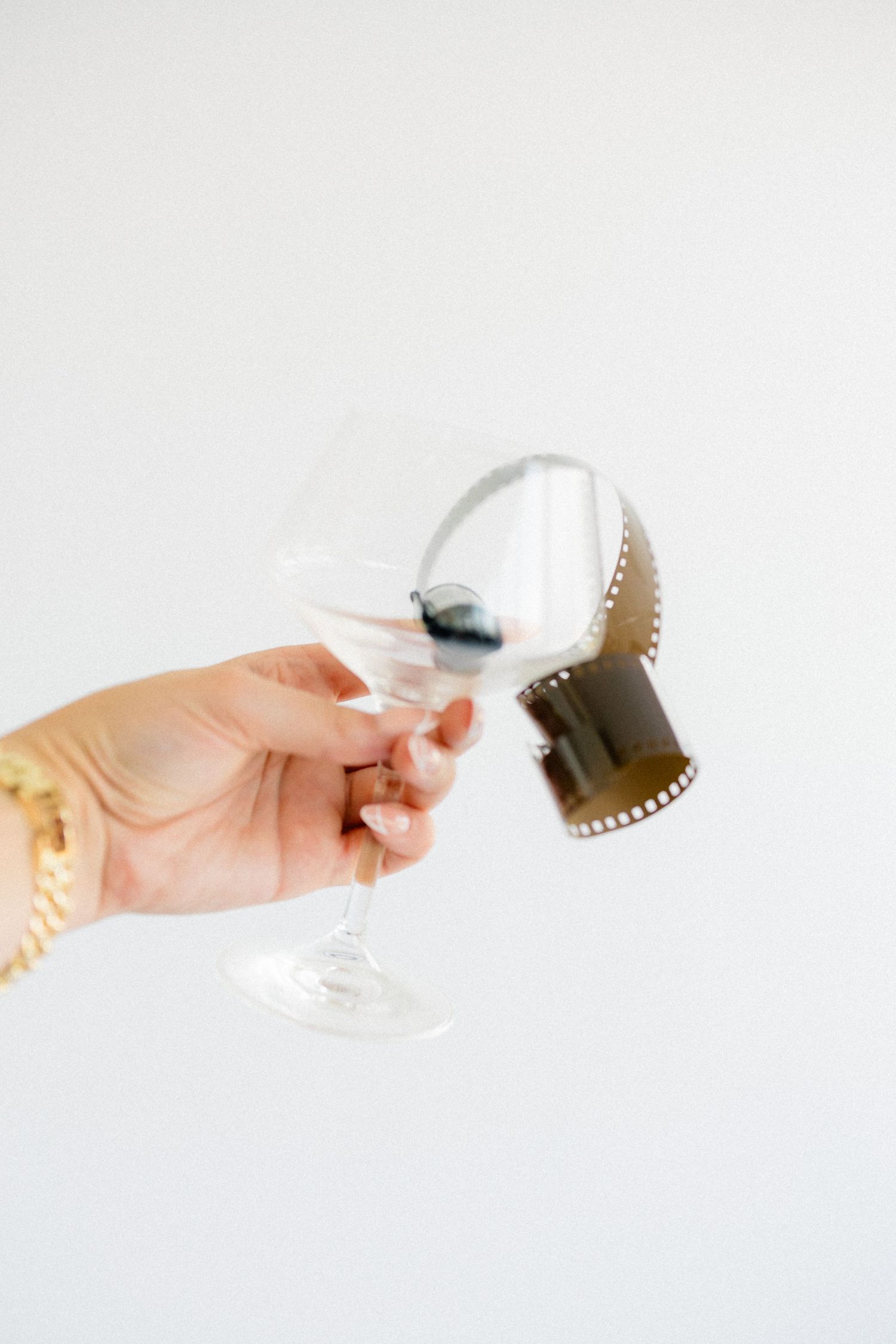 Hand holding a clear wine glass containing a roll of 35mm film strip against a white background.