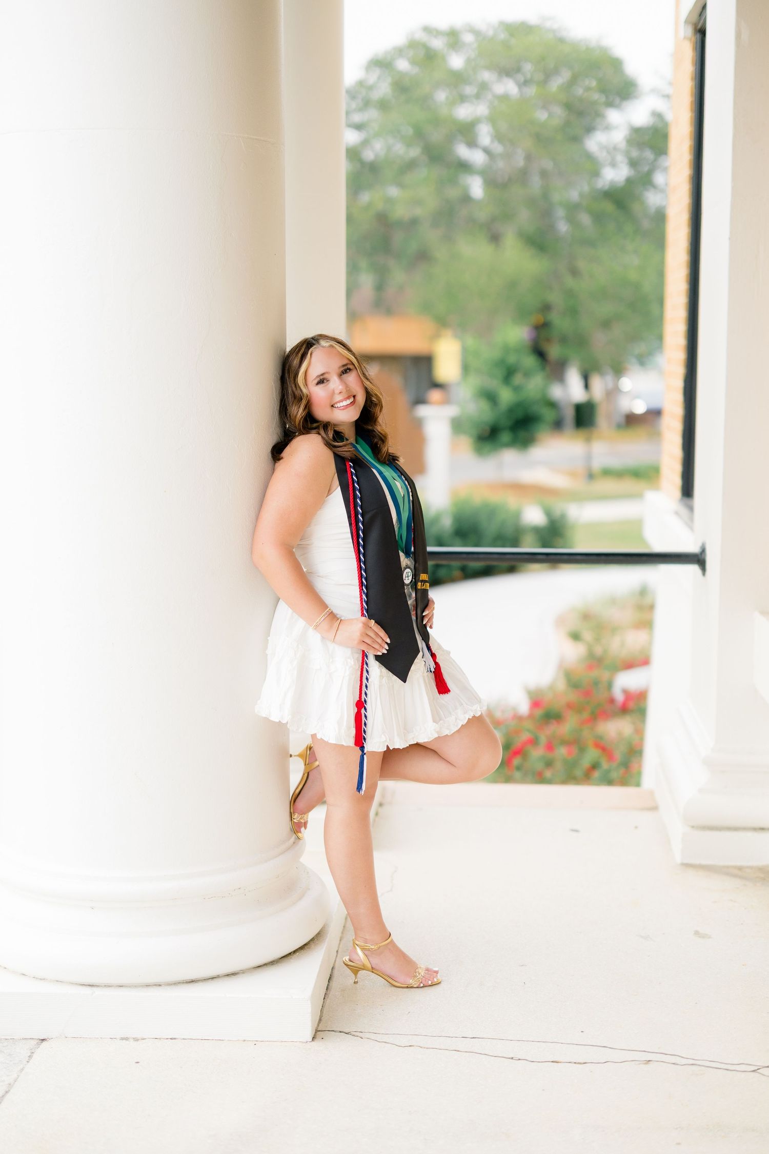 Smiling graduate in white dress with academic stoles leans against white column outdoors on a sunny day.