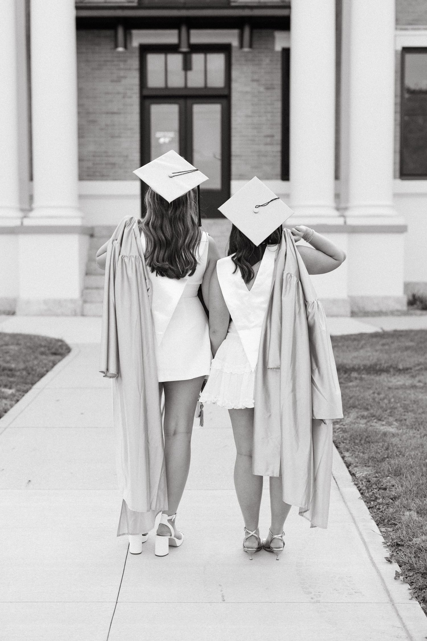 Two female graduates walking away in caps and gowns, wearing white dresses, on a campus sidewalk, black and white photo.