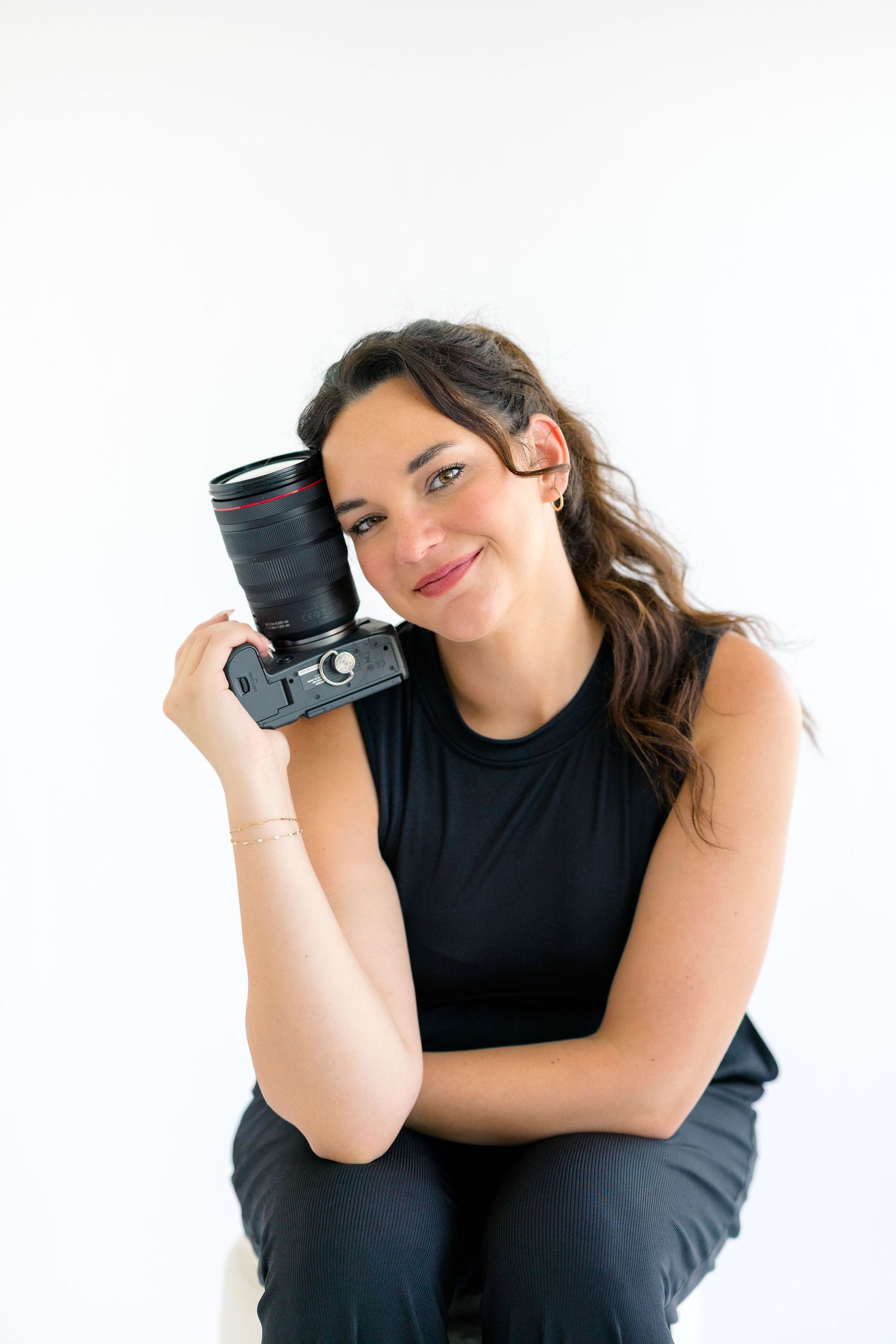 Smiling woman in black outfit holding a DSLR camera against white background, perfect for photography business profiles.