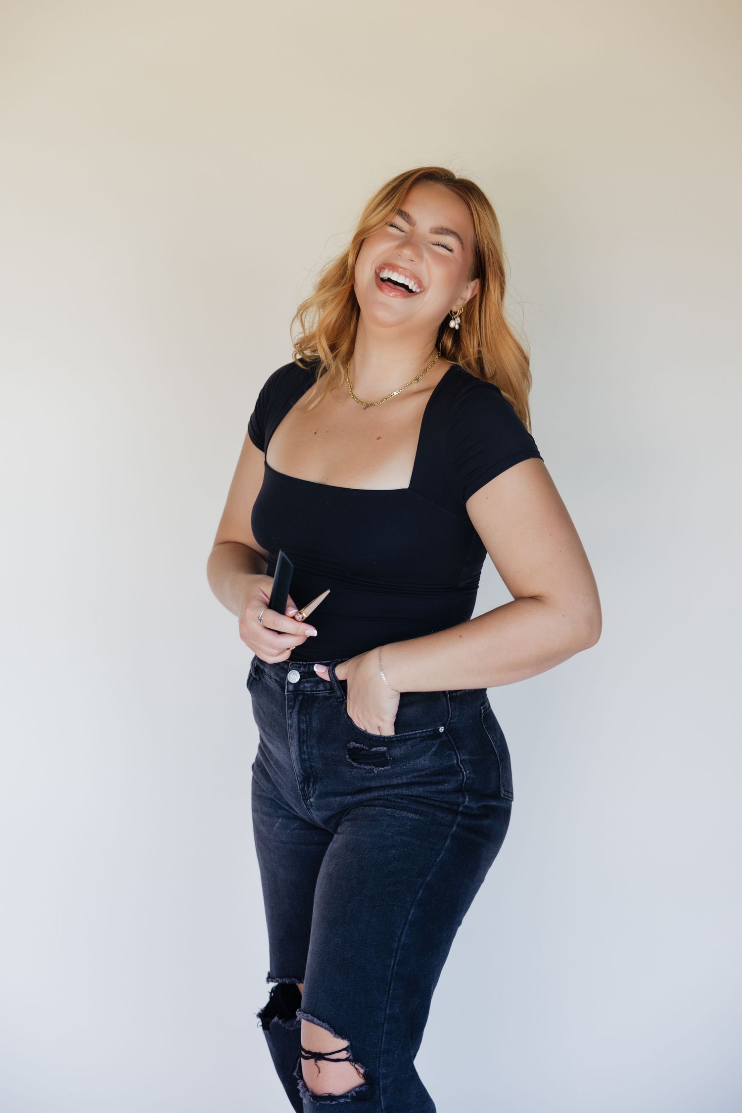 Woman in black top and ripped jeans laughing joyfully against white background.