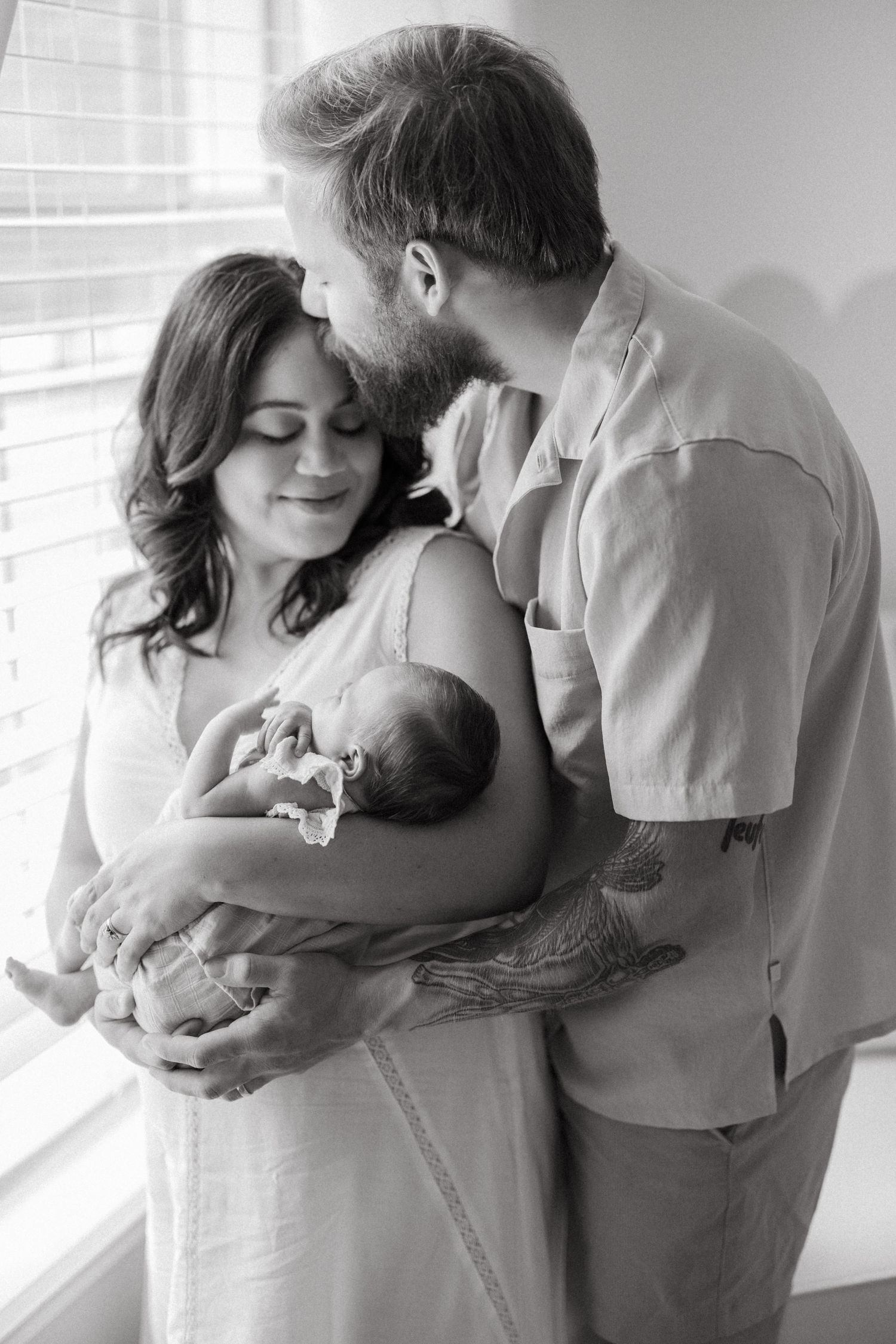 Black and white photo of new parents tenderly holding their newborn baby by a window.